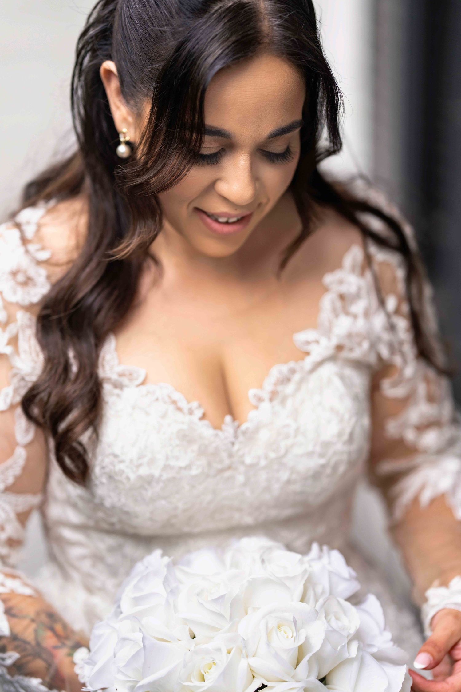 Dark-haired bride in lace long-sleeve wedding gown holding white rose bouquet, looking down softly.