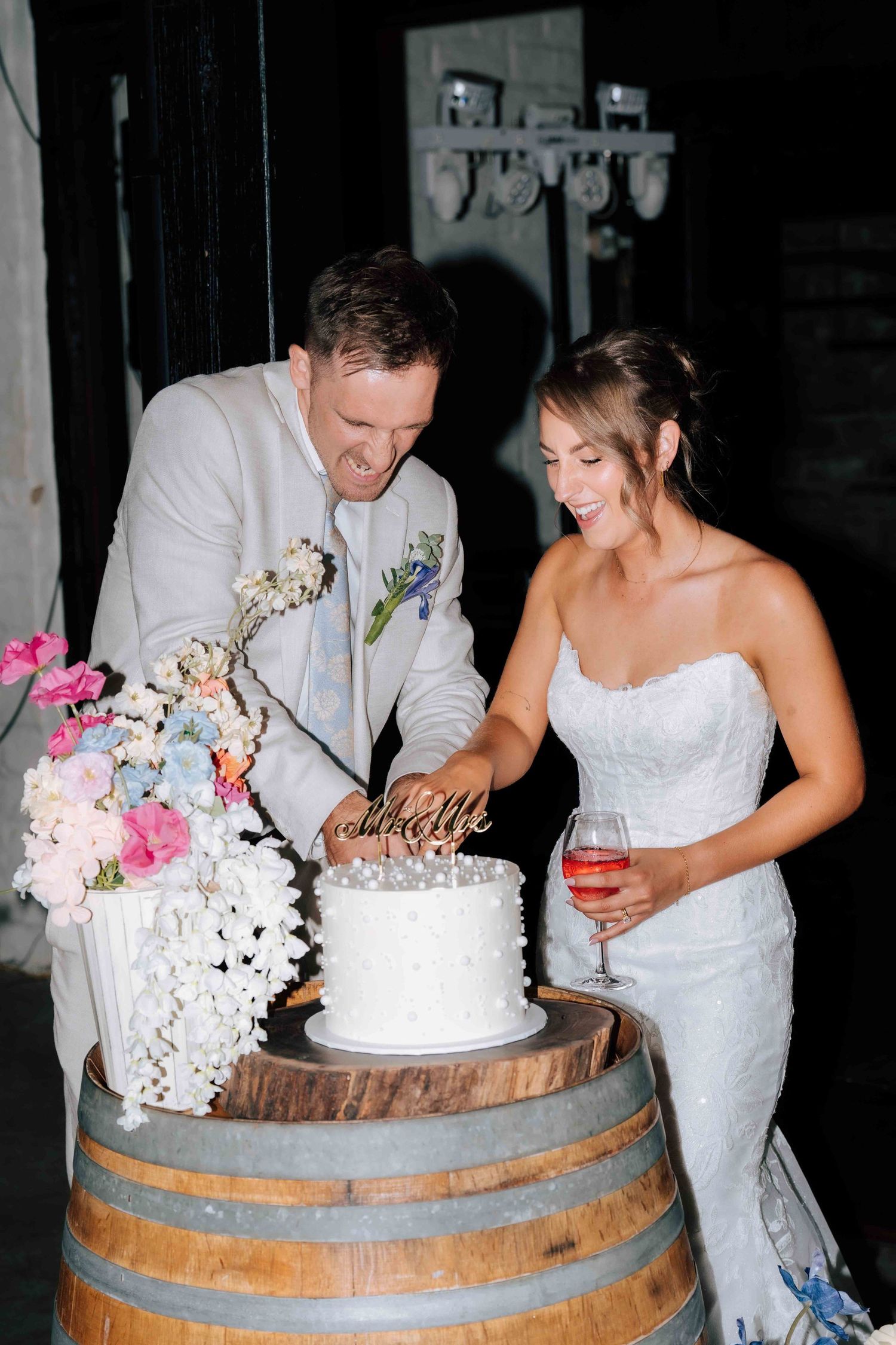 Bride and groom laughing while cutting white wedding cake on barrel, with colorful floral arrangement nearby.