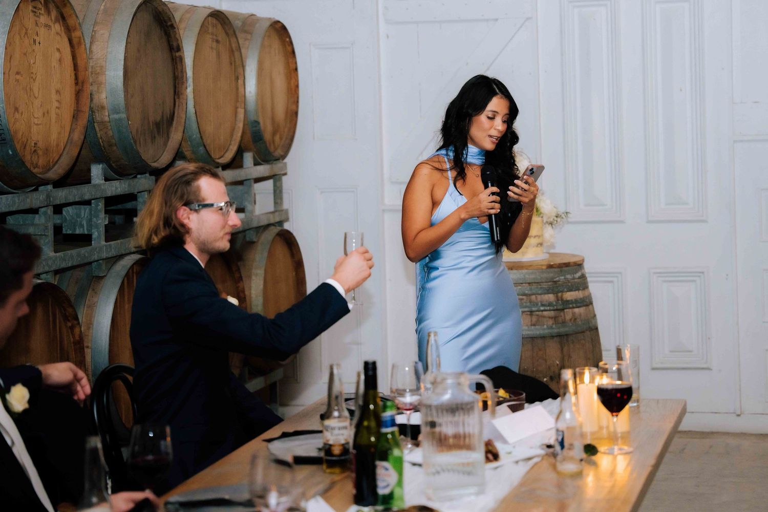 Bridesmaid in blue dress gives a speech holding a microphone beside wine barrels at a winery wedding reception.