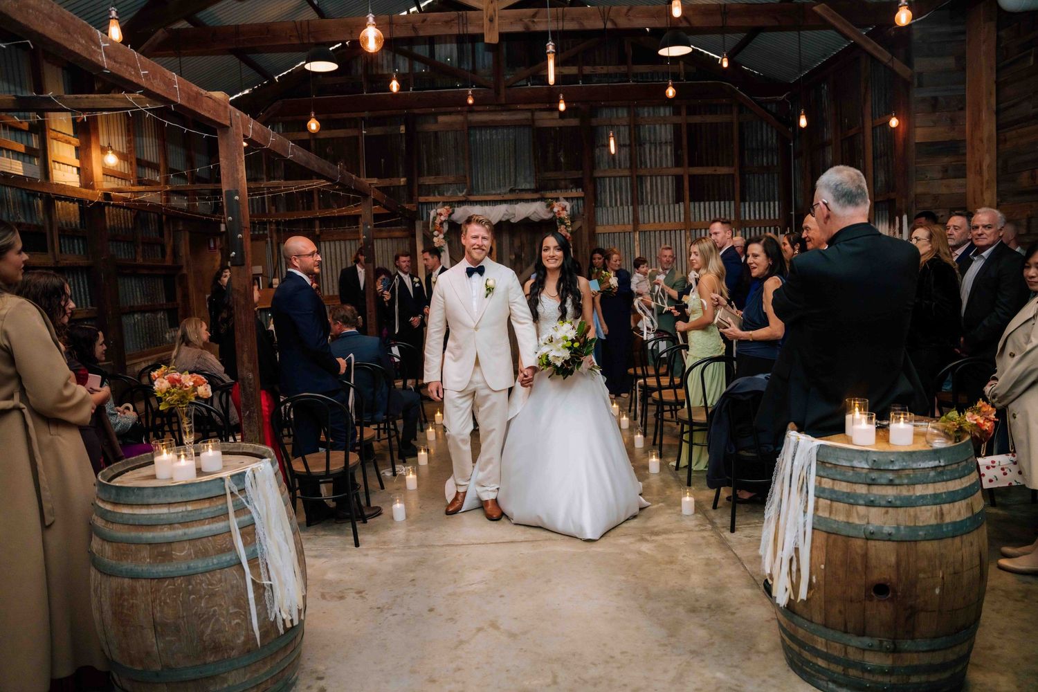 Bride and groom walk down the aisle in a rustic barn wedding venue with barrel decorations and candlelight.