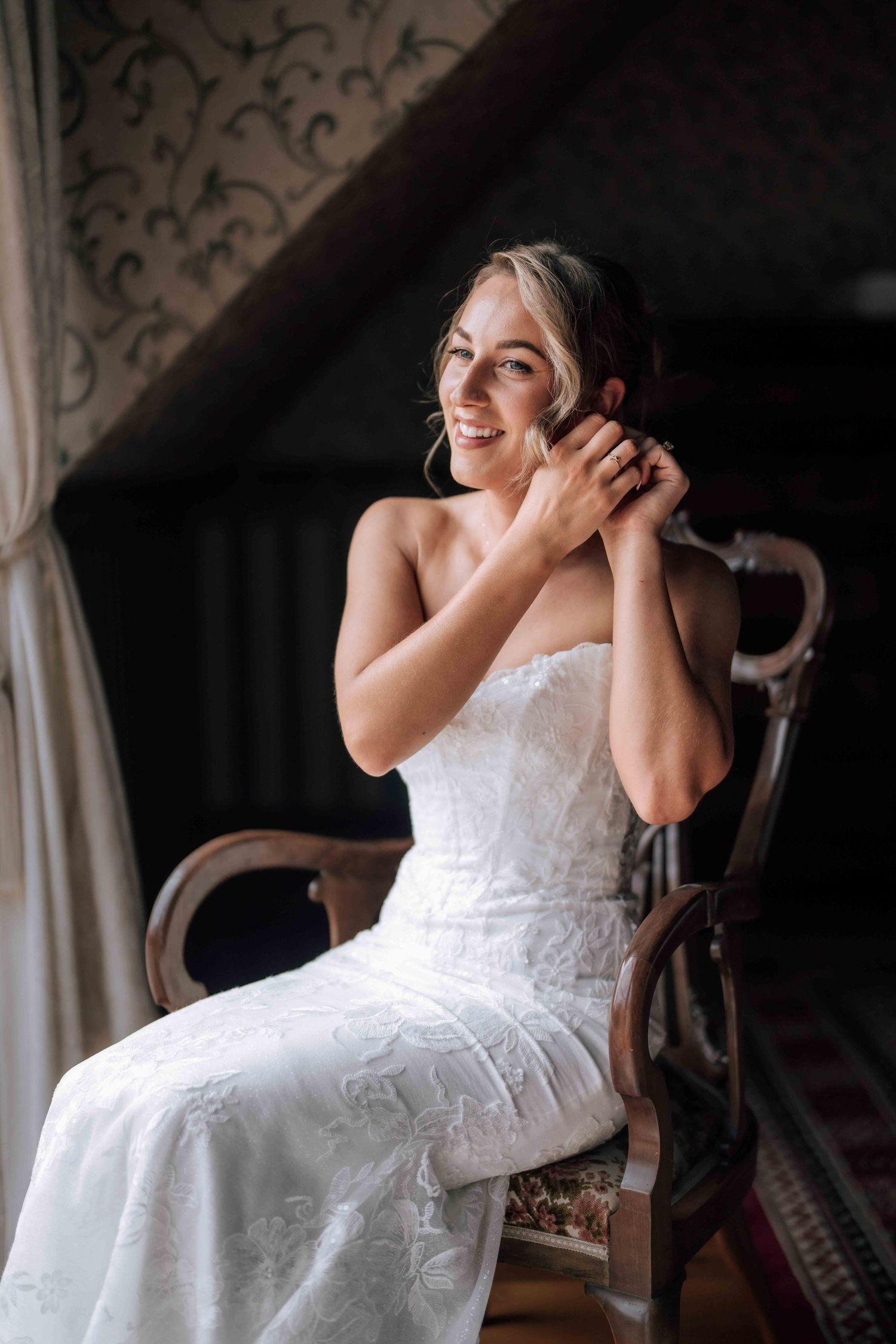 Radiant bride in strapless lace wedding dress adjusting earring while seated in wooden chair.