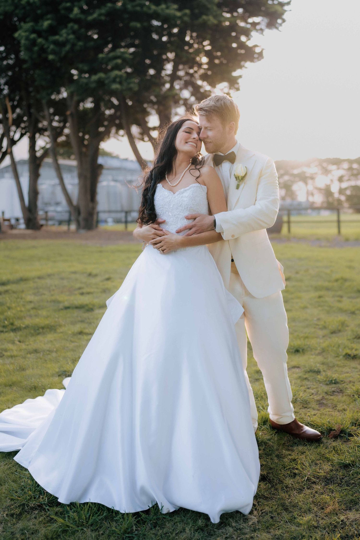 Bride in white gown and groom in cream suit embrace at golden hour in a lush green park.