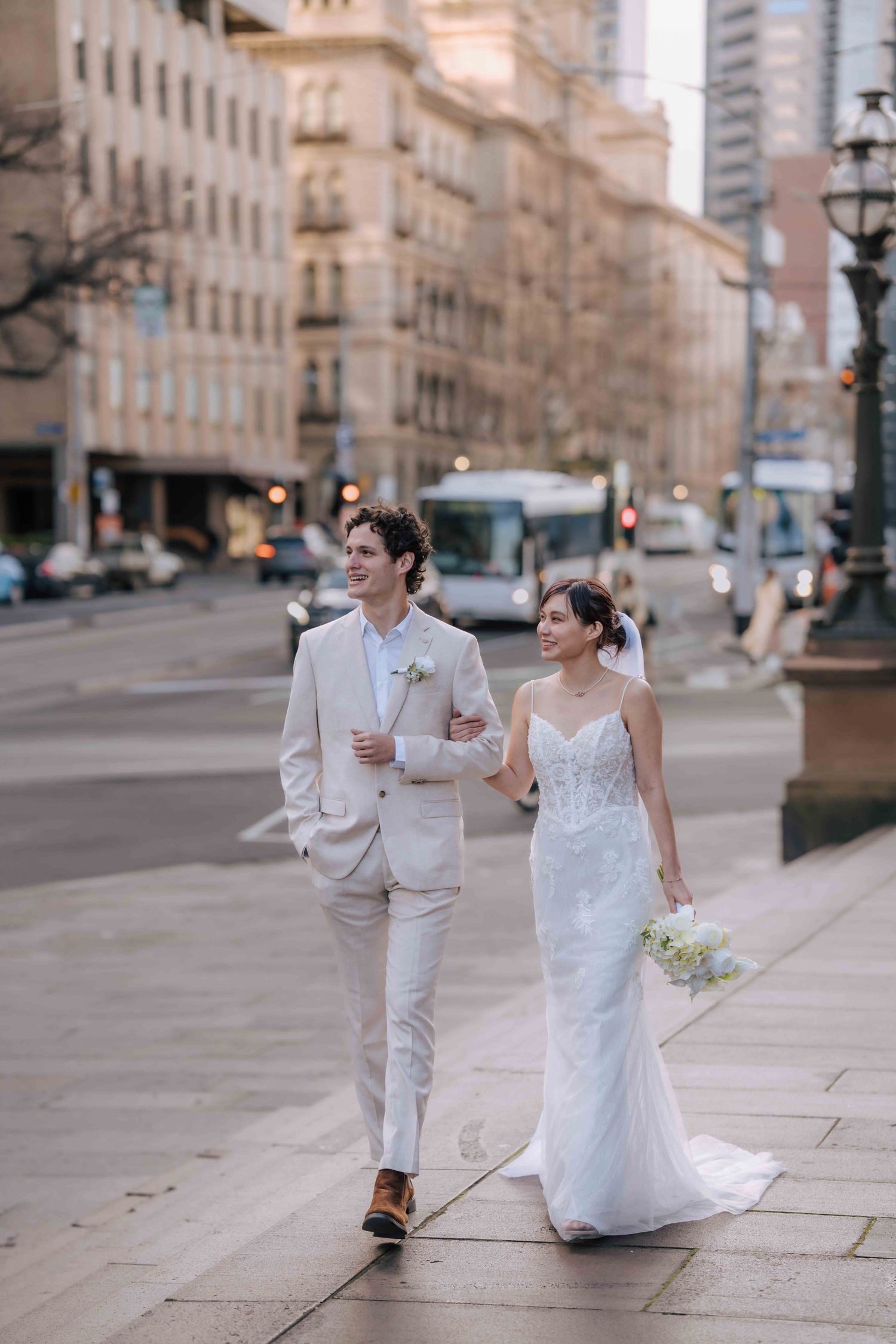 Bride and groom walk arm in arm along city street, she in white gown, he in beige suit.