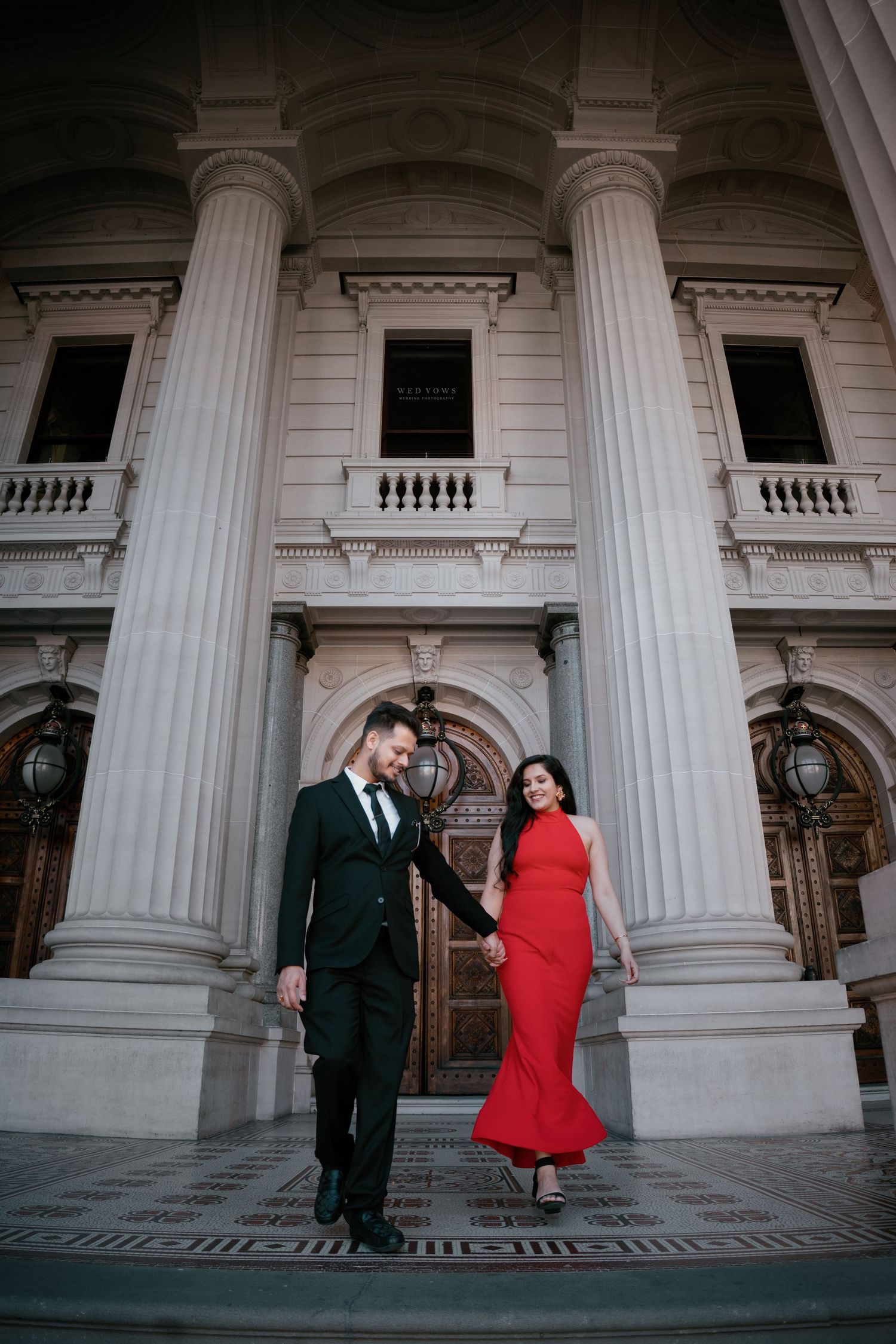 Elegant couple in red dress and black suit holding hands before grand neoclassical building columns.