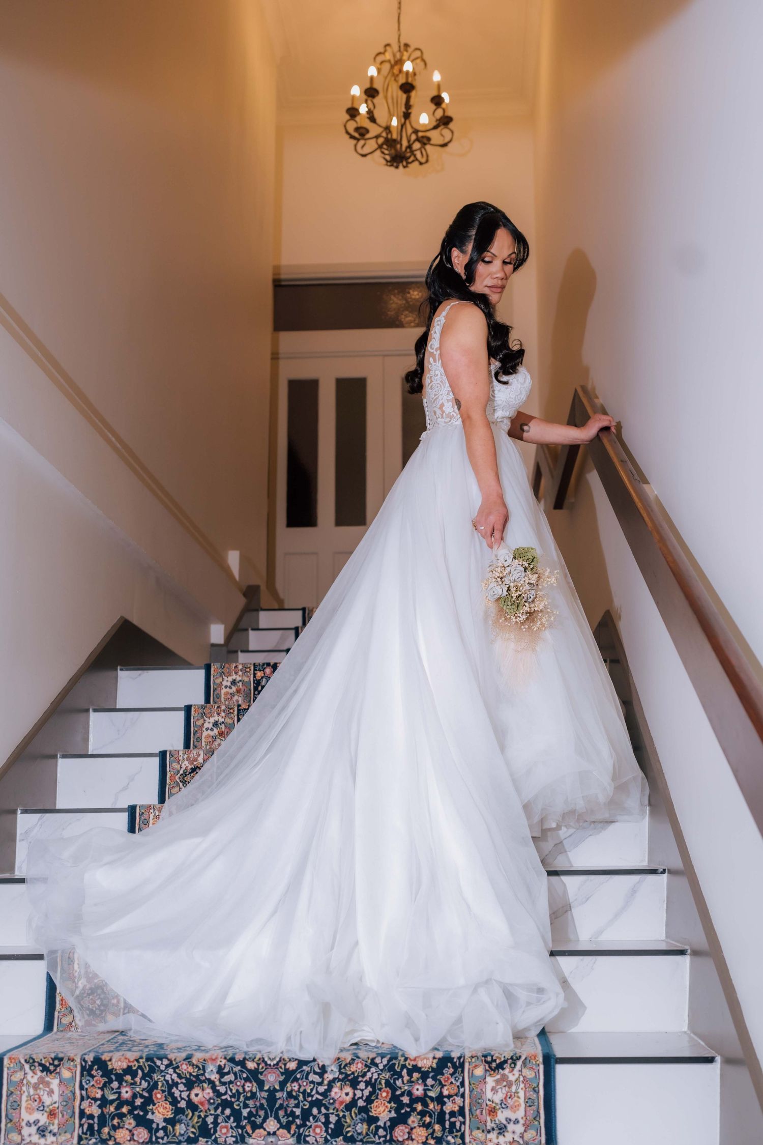 Bride in a white lace ball gown descending a staircase while holding a bouquet beneath a chandelier.
