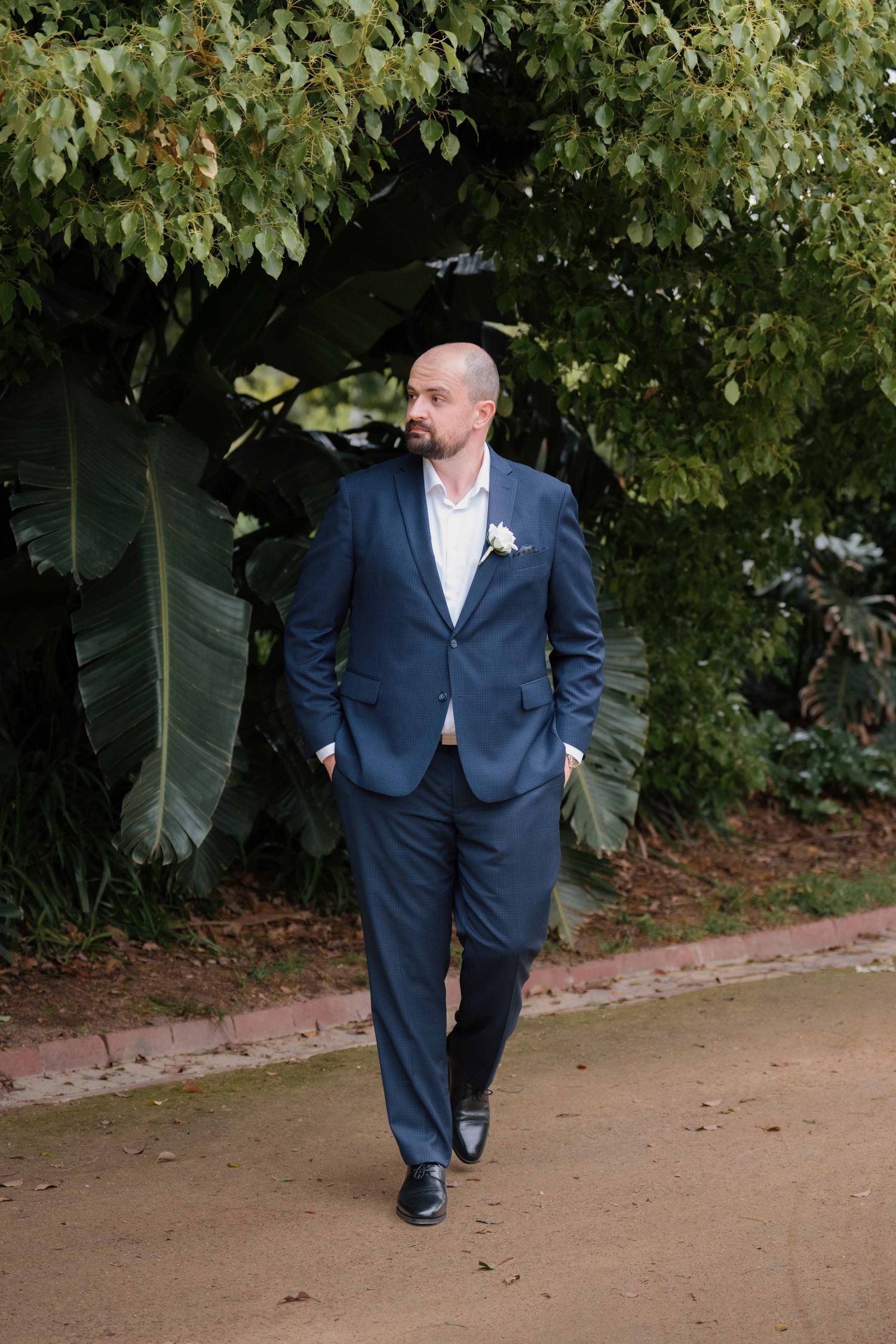Groom in navy blue suit with white boutonniere walking along garden path surrounded by lush green tropical foliage.
