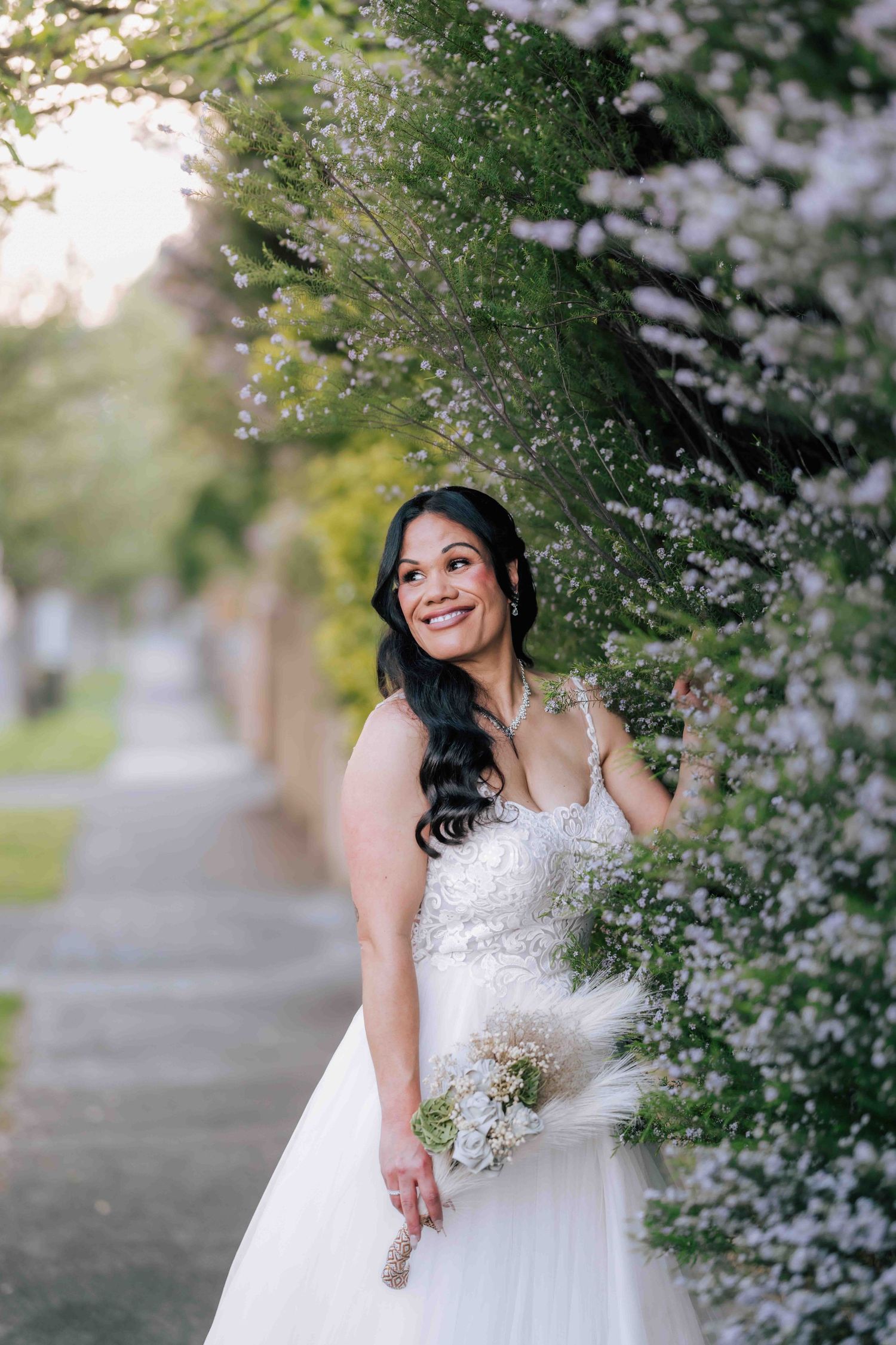 Joyful bride in beaded gown smiling among delicate white flowering outdoor trees.