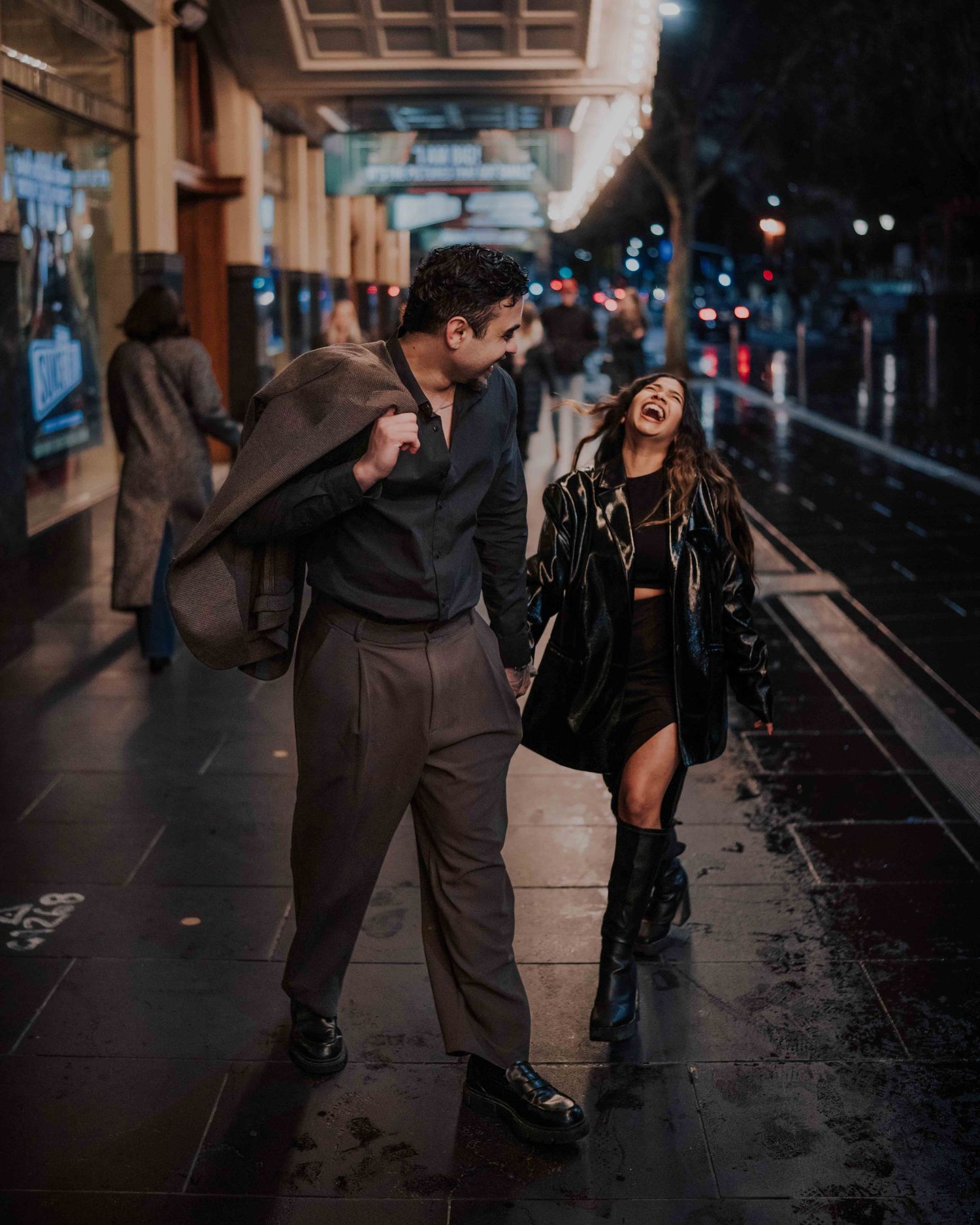 Couple laughing and holding hands on a rainy city street at night, woman in black leather jacket.