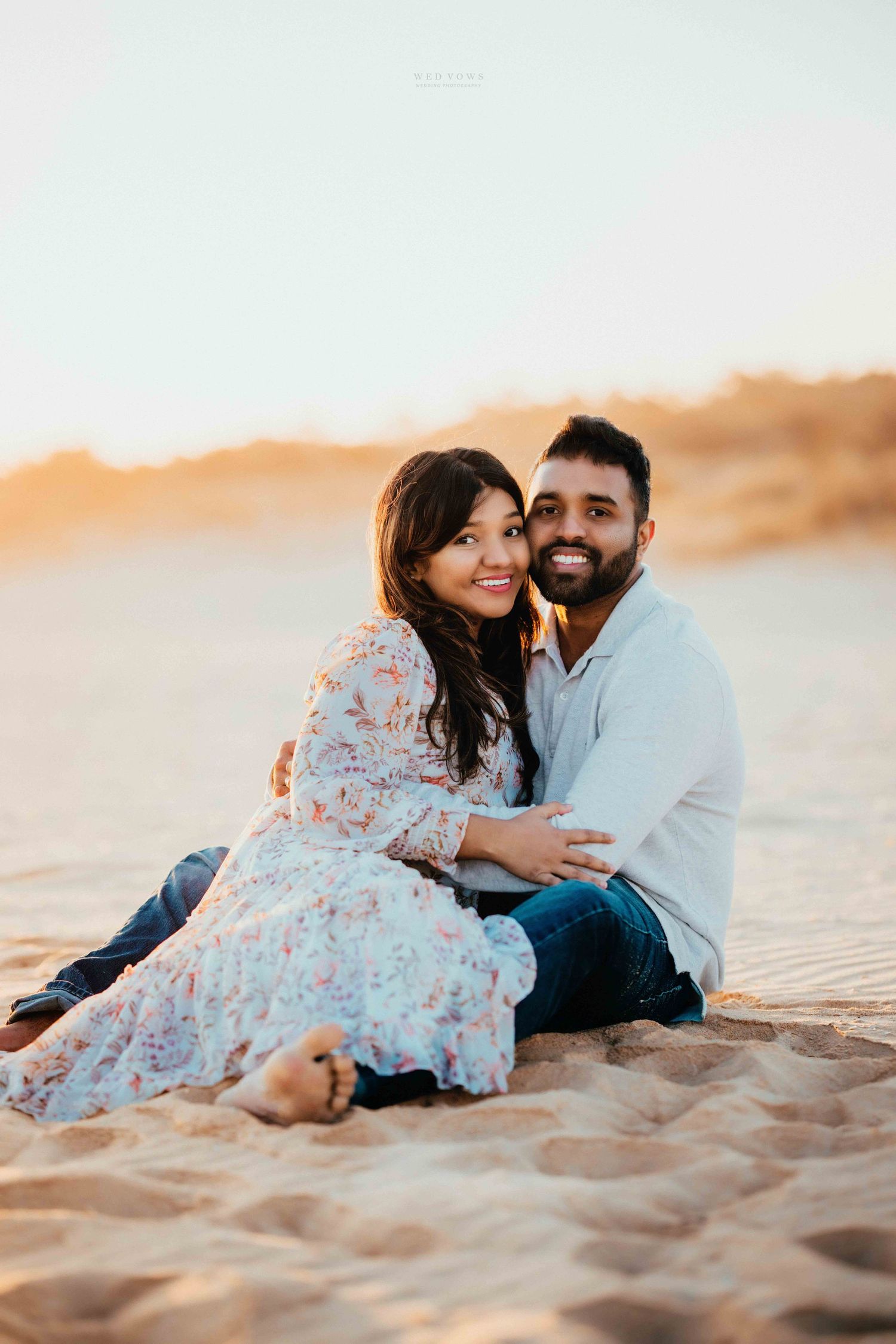 Happy couple sitting together on sandy beach smiling at camera during golden hour sunset.