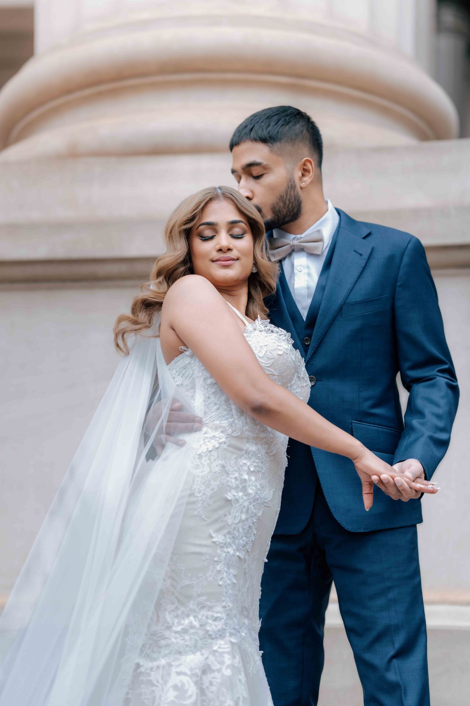 Groom in navy suit kissing bride's temple while holding hands near elegant stone columns.