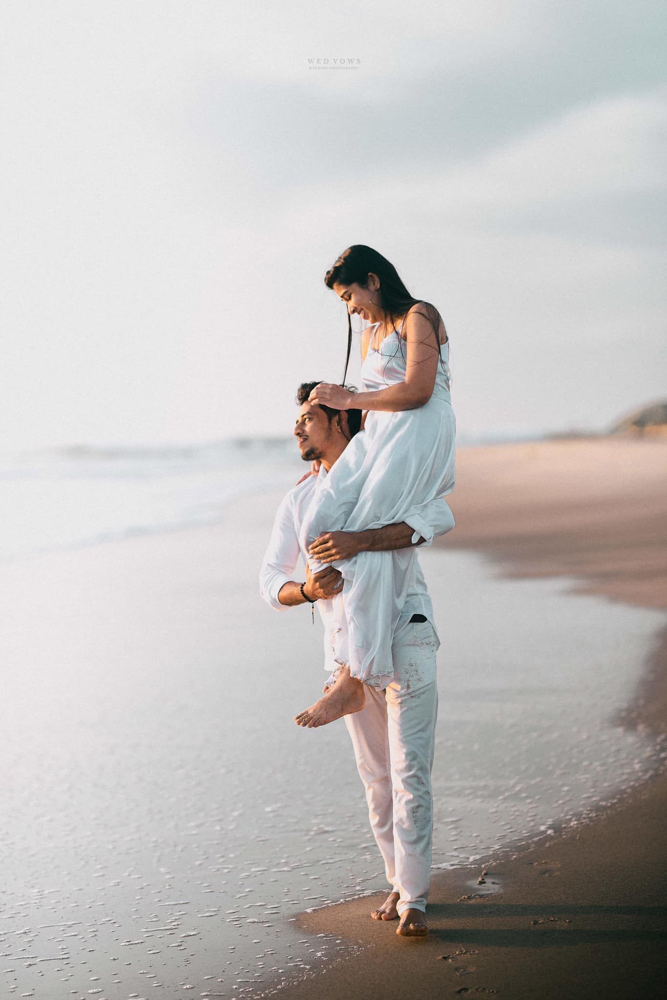 Man carrying woman on shoulders while walking barefoot on sandy beach at sunset.