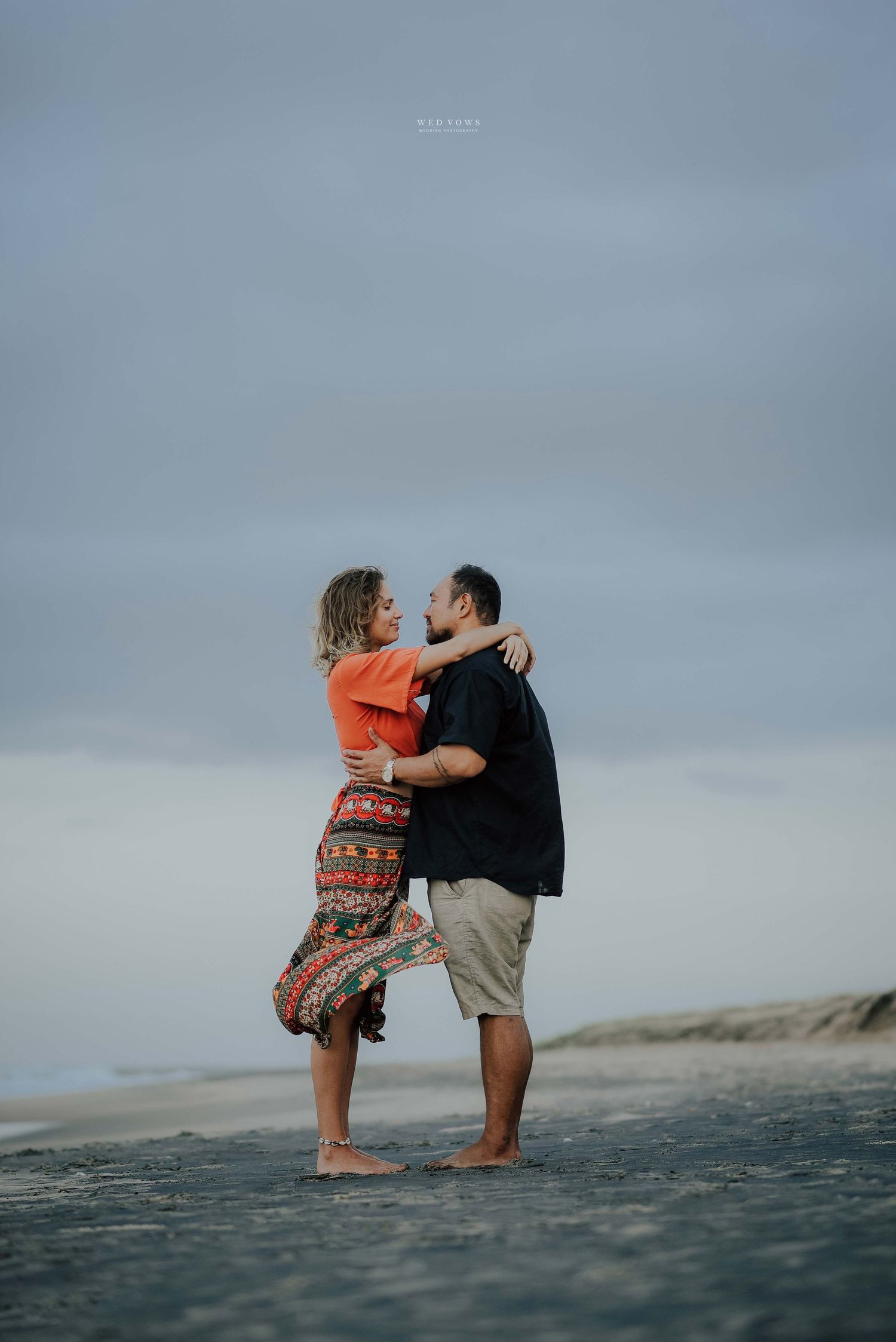 A couple shares a romantic almost-kiss moment while standing on a windswept beach under a cloudy sky.