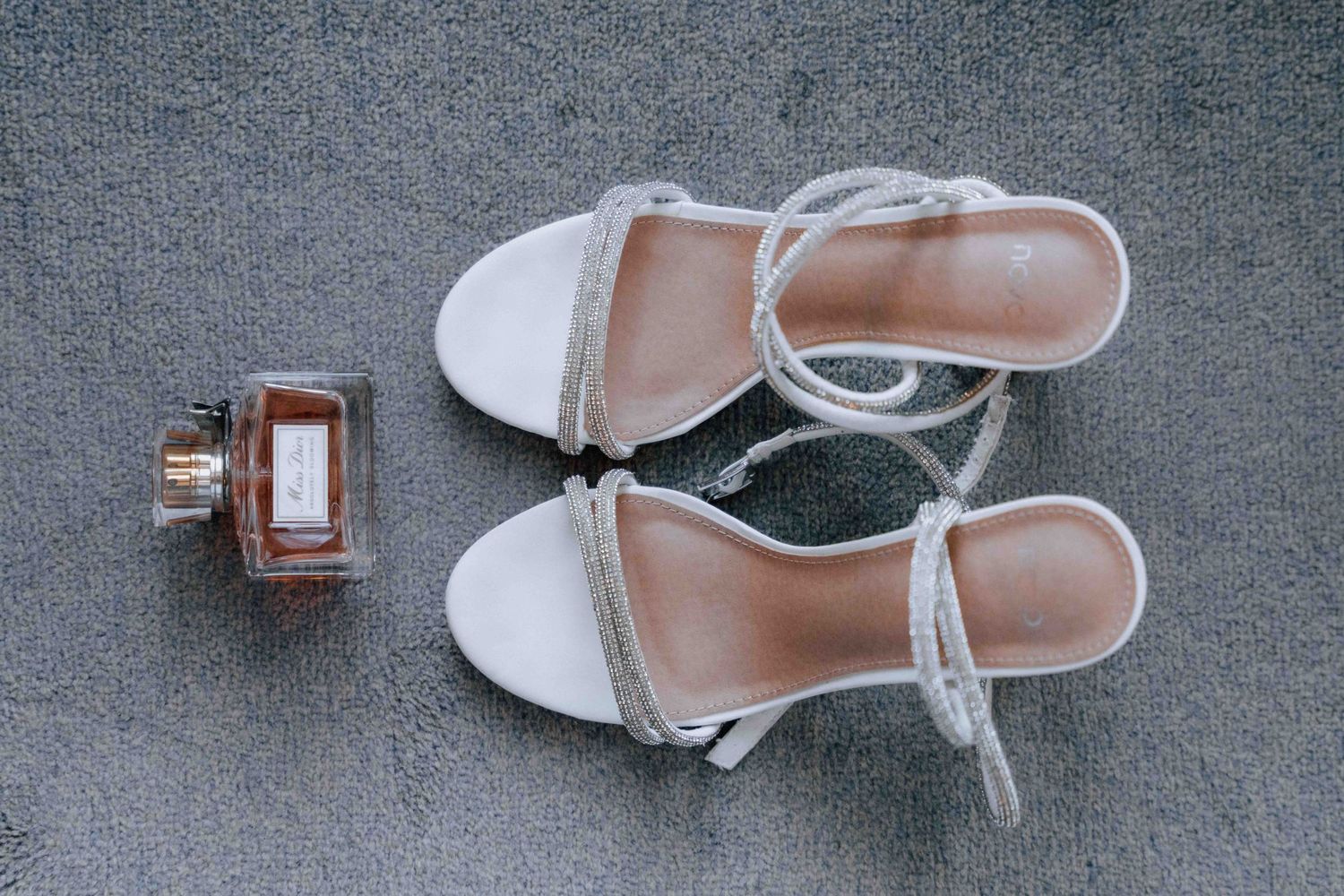 Top view of white strappy bridal heels with rhinestone details beside amber perfume bottle on carpet.