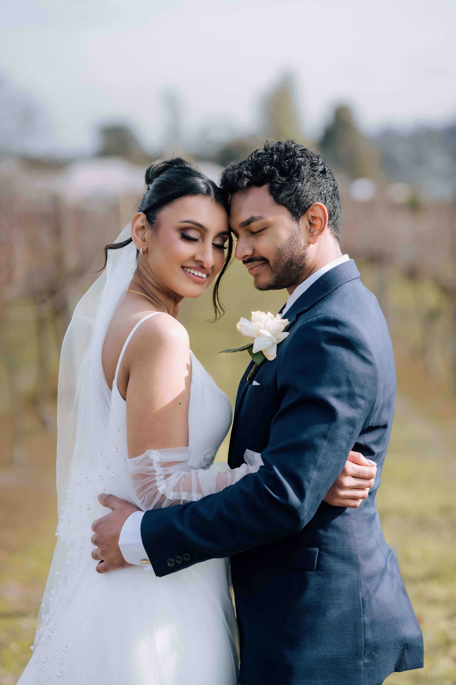 Smiling bride and groom embrace closely in a vineyard, groom wearing white rose boutonniere.