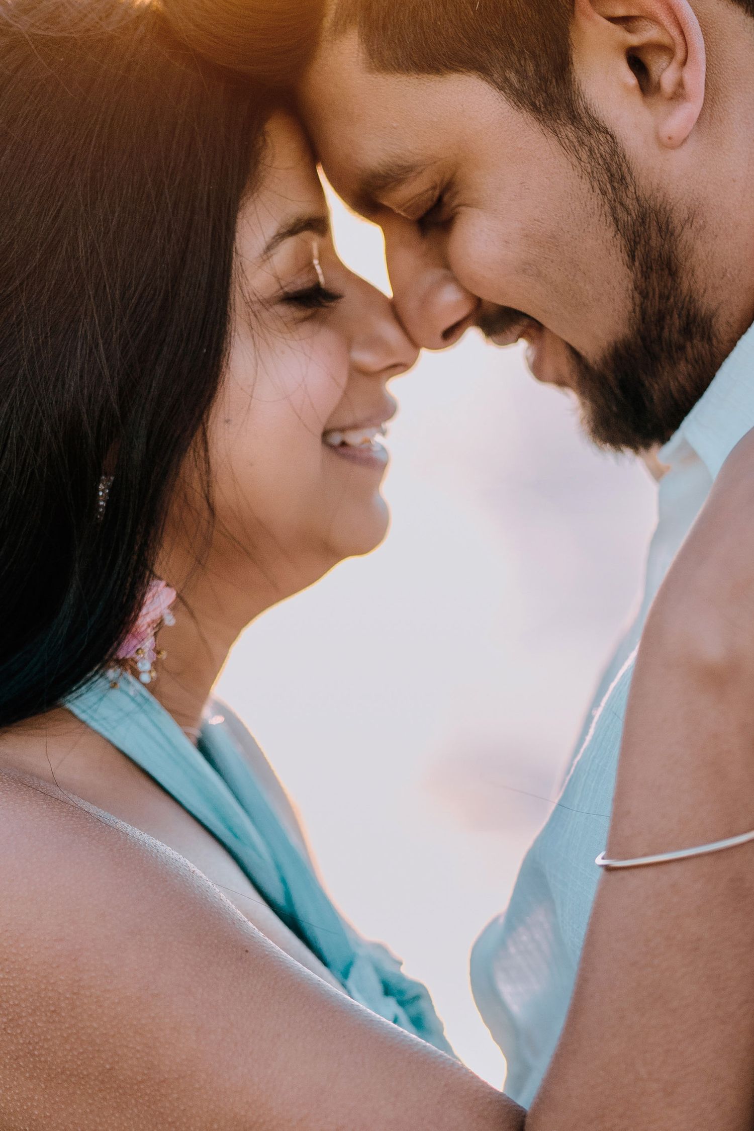 Close-up of smiling couple touching foreheads tenderly, woman with pink earrings in soft golden light.