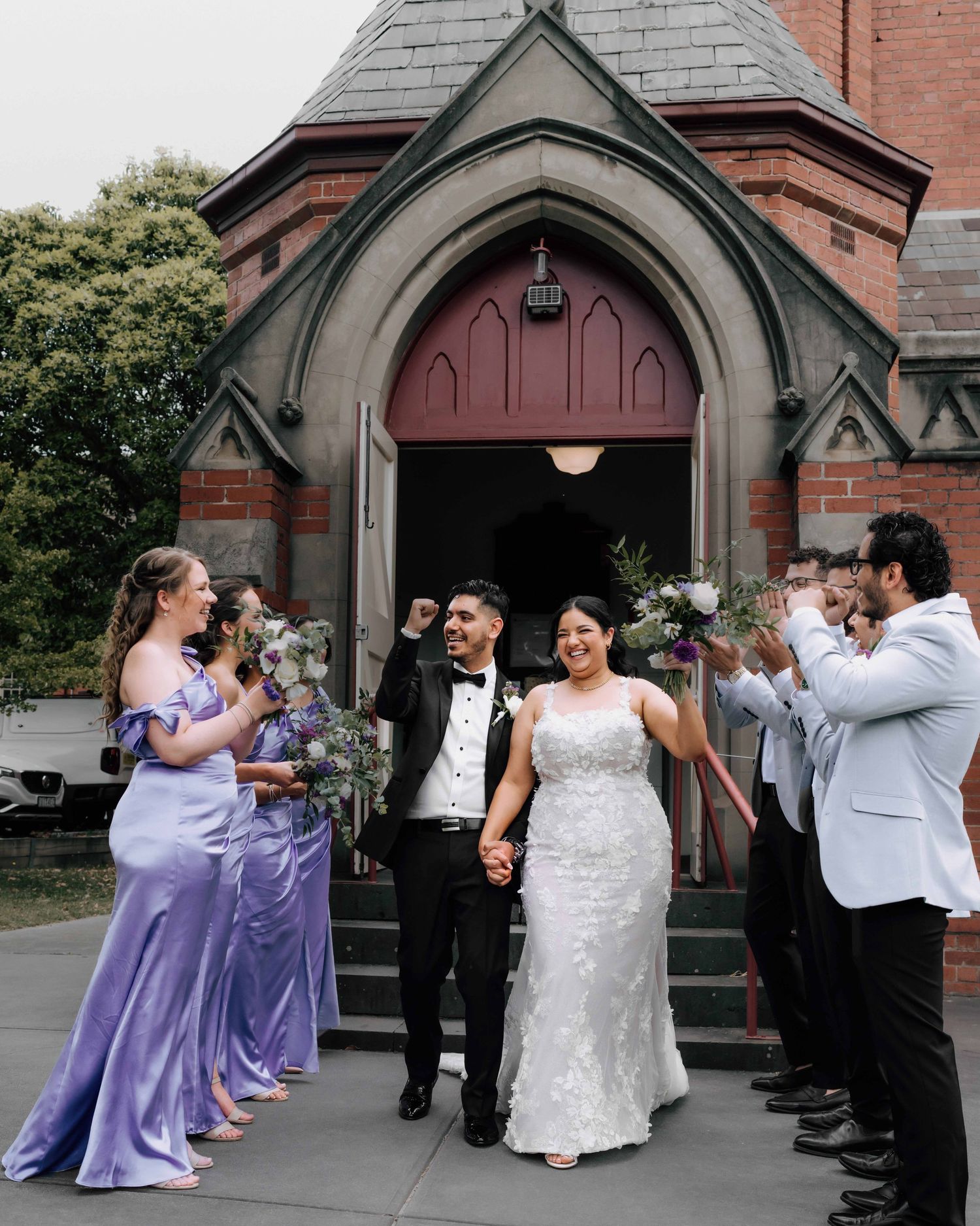 Jubilant newlyweds exit red brick church as bridesmaids in lavender and groomsmen in white cheer.
