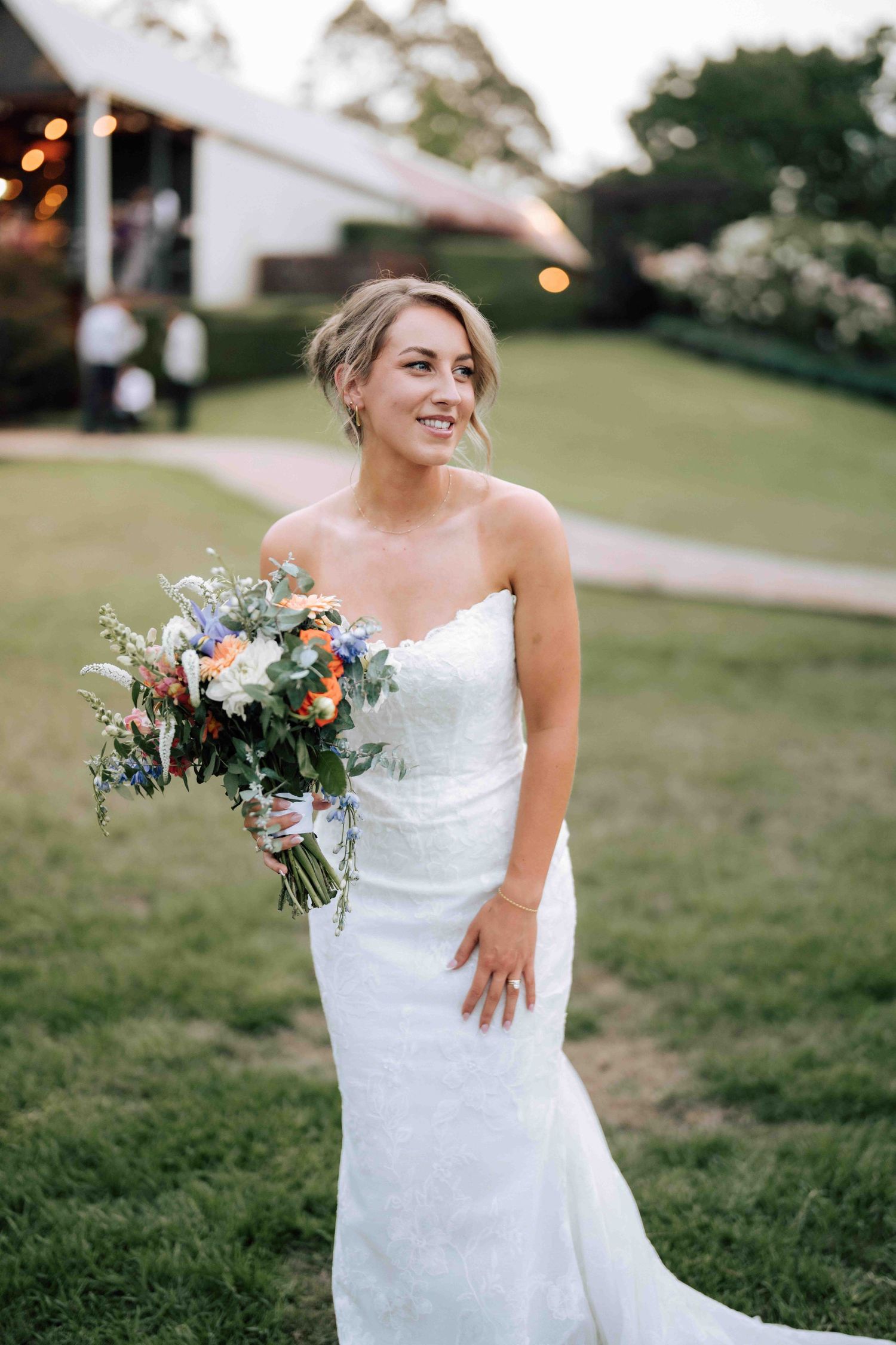 Bride in strapless lace wedding gown holding colorful bouquet smiling outdoors at garden venue.