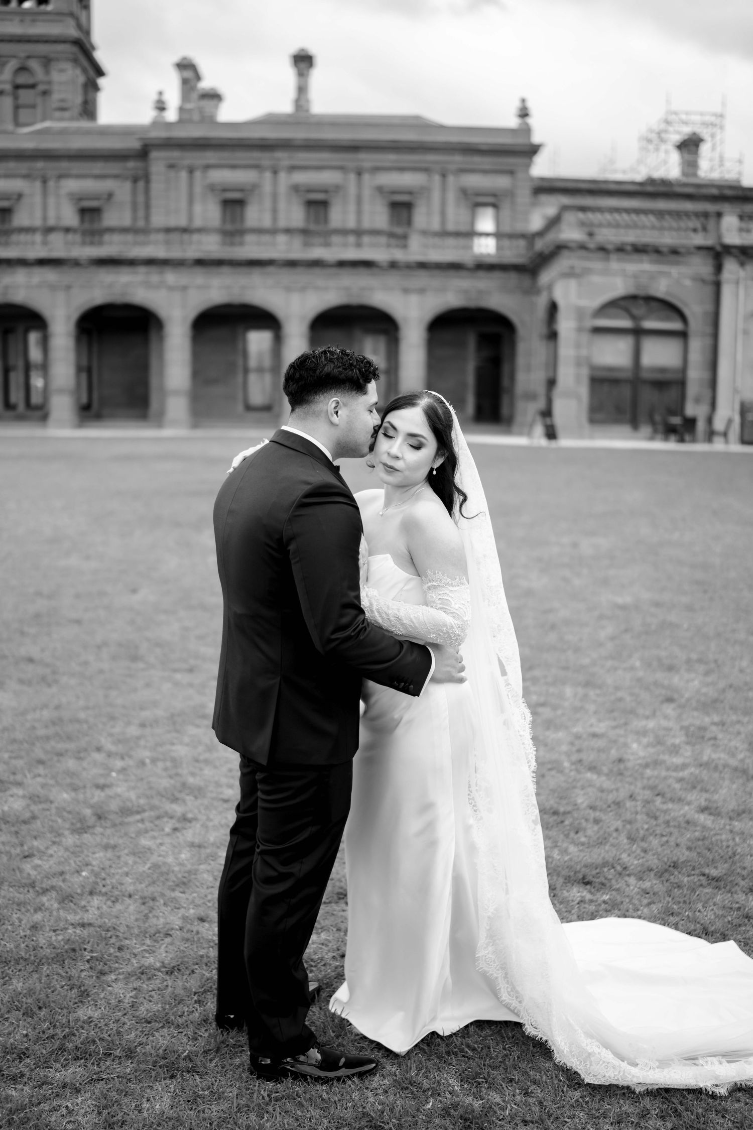 Bride and groom share a romantic embrace on lawn in black and white portrait before historic building.