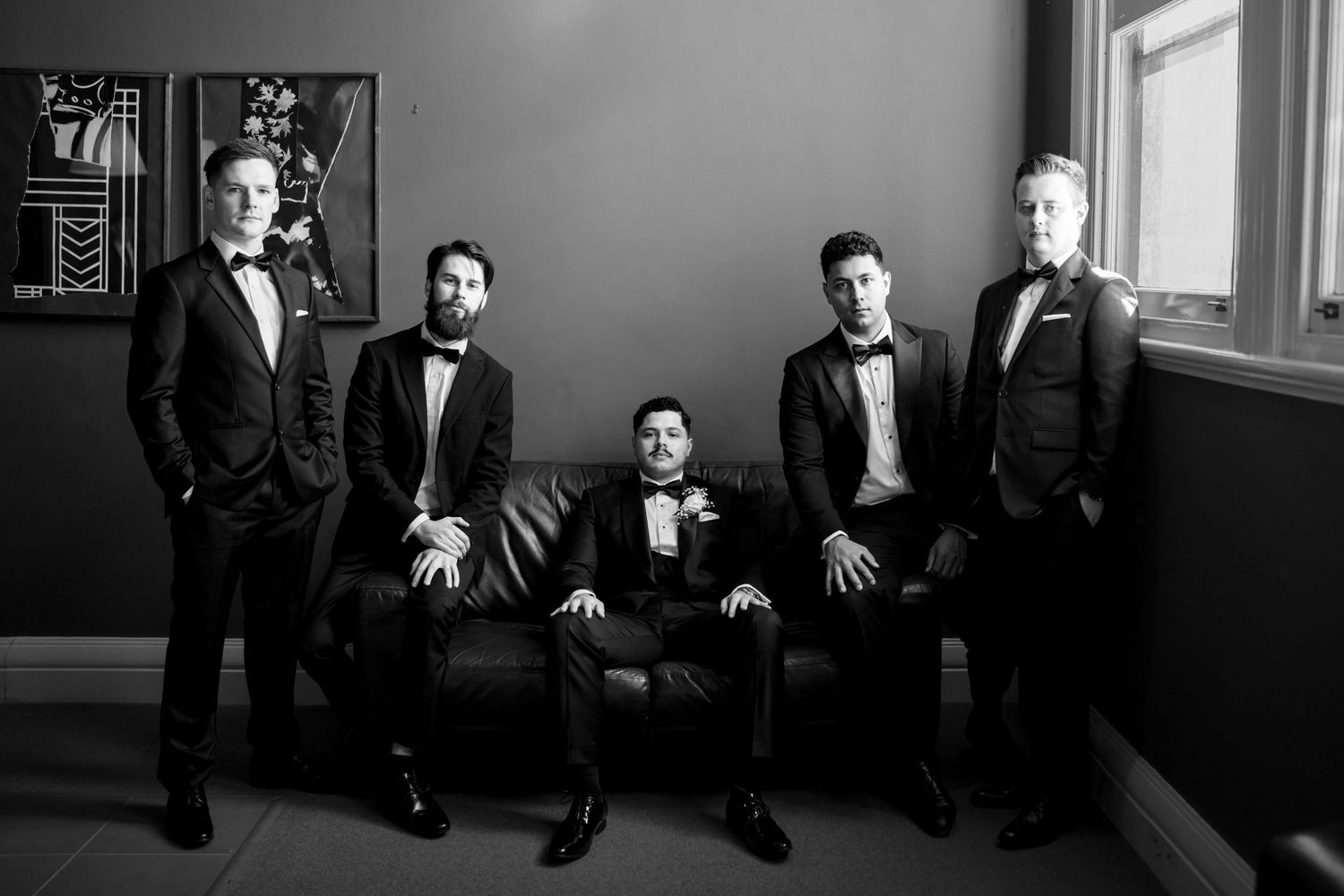 Five men in black tuxedos pose formally in a black and white photo, one seated on a leather sofa.
