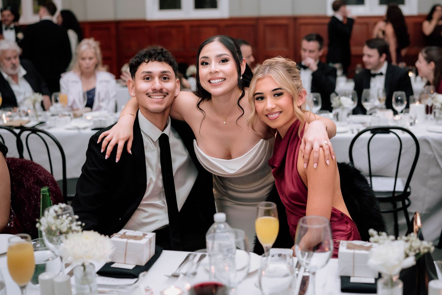 Three smiling wedding guests posing at reception dinner table with floral centerpieces and champagne.