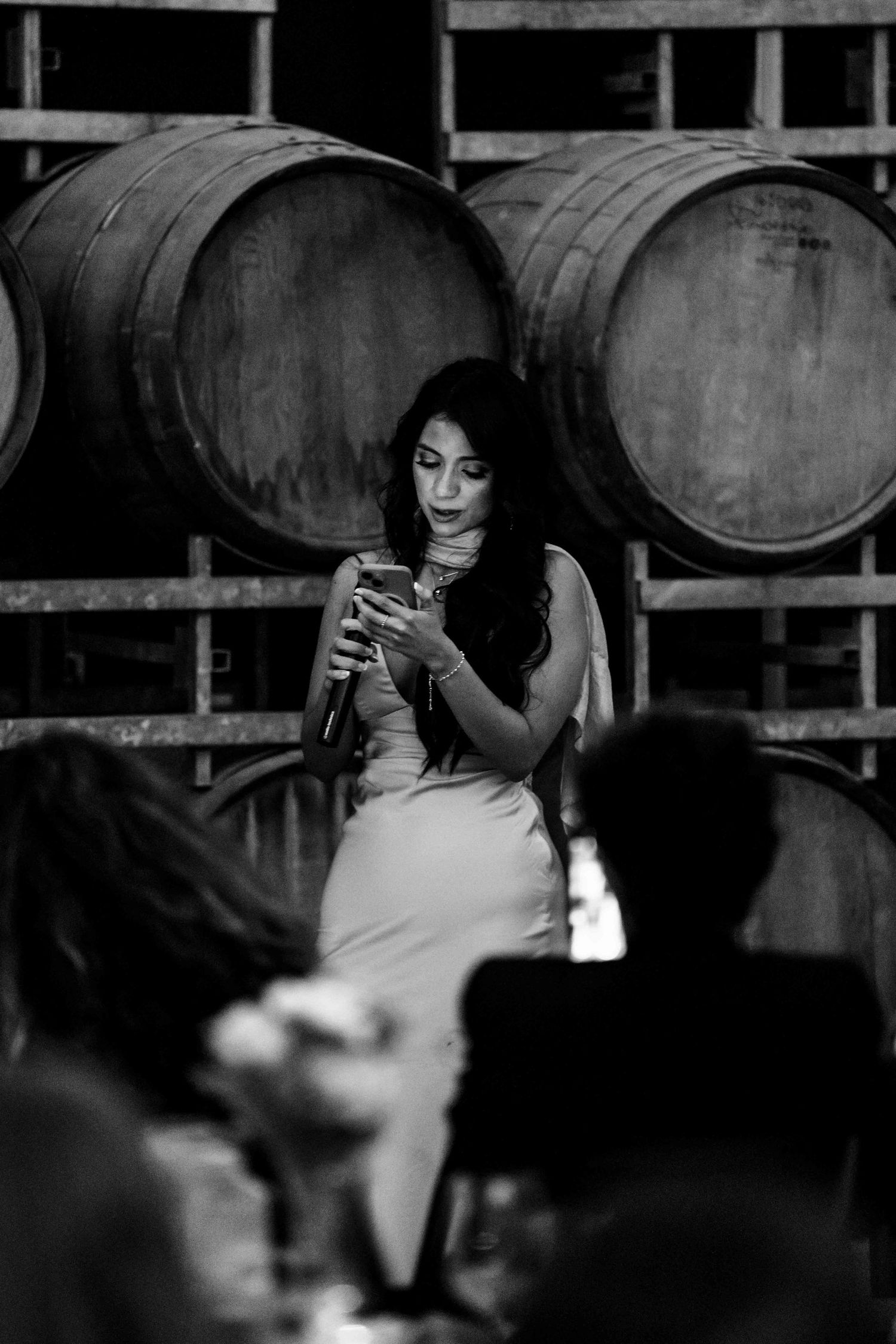 Woman in elegant dress reads from phone near wine barrels at winery wedding reception, black and white photo.