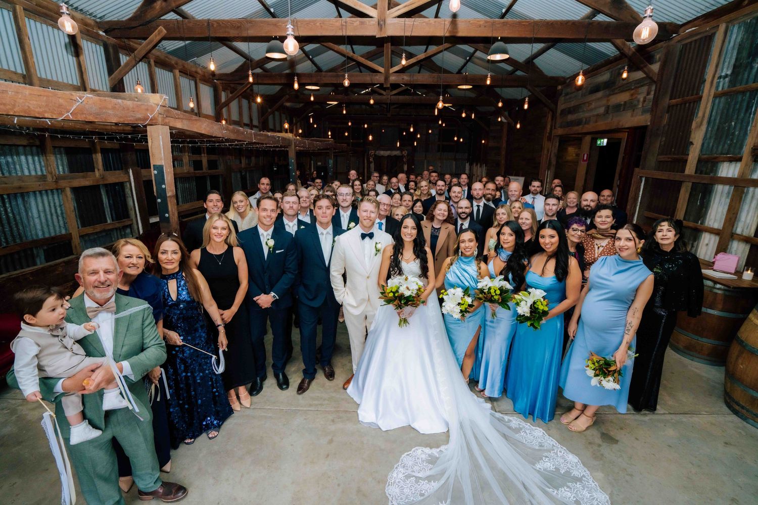 Large wedding group photo with bride in white gown and groom in white tuxedo surrounded by guests in a rustic barn venue.