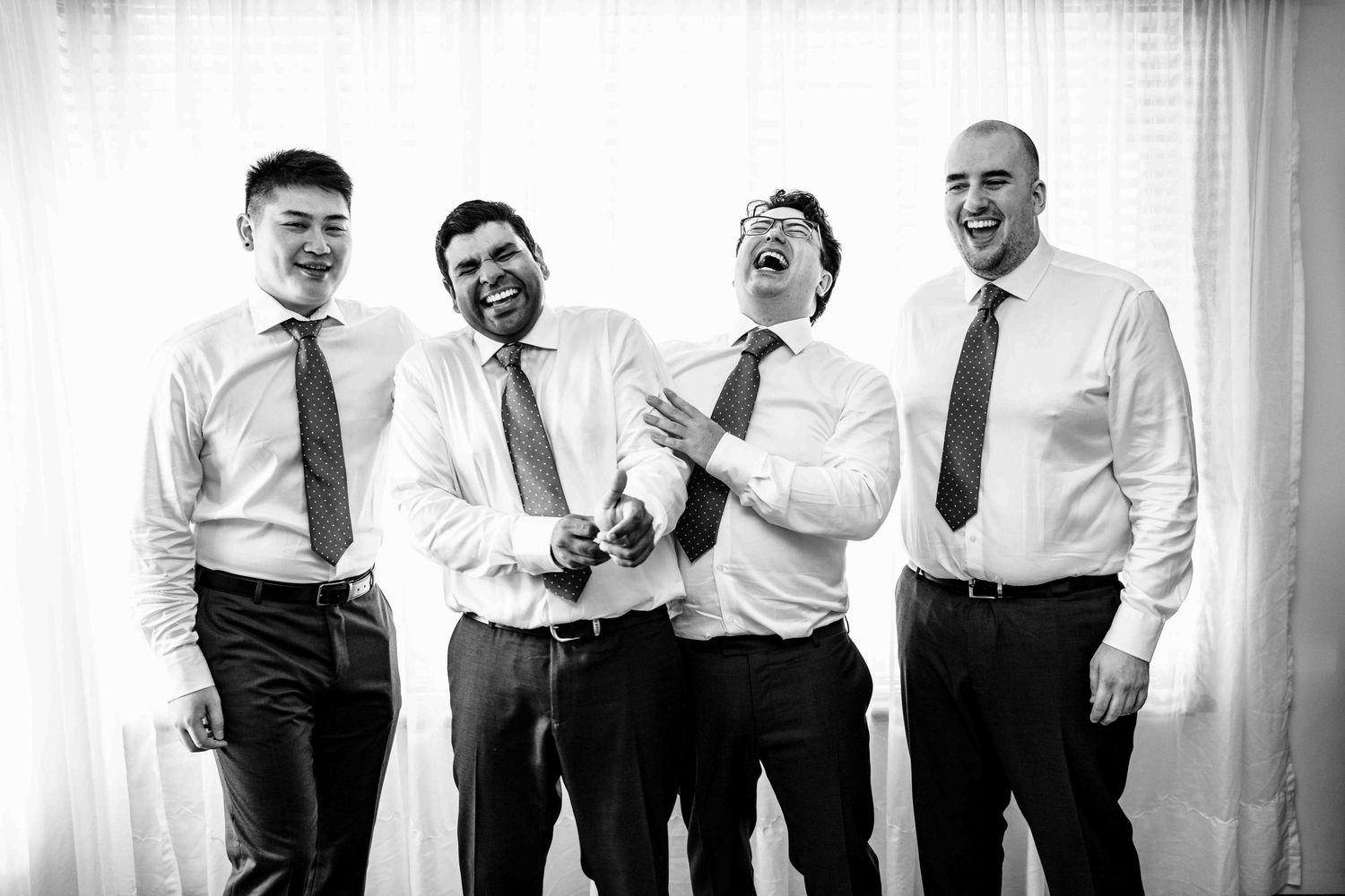 Four groomsmen in white shirts and ties laugh together in black and white pre-wedding portrait.