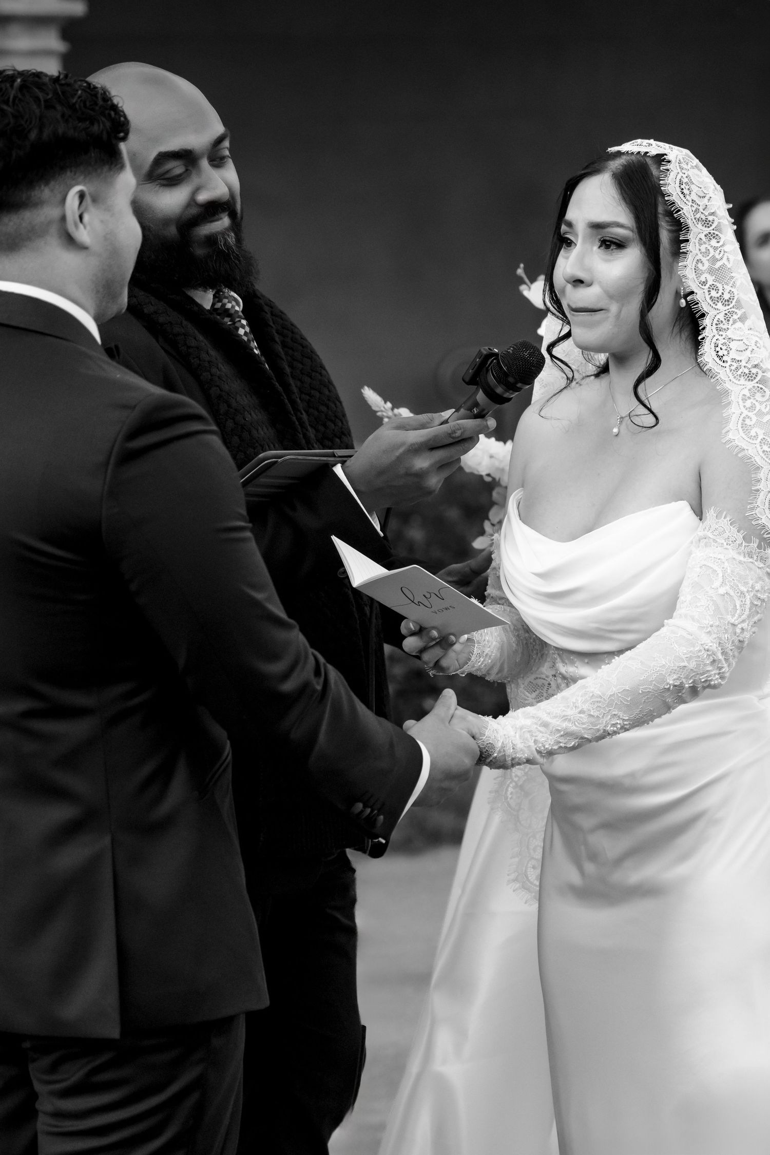 Bride and groom exchange vows and rings with officiant in emotional black and white ceremony portrait.