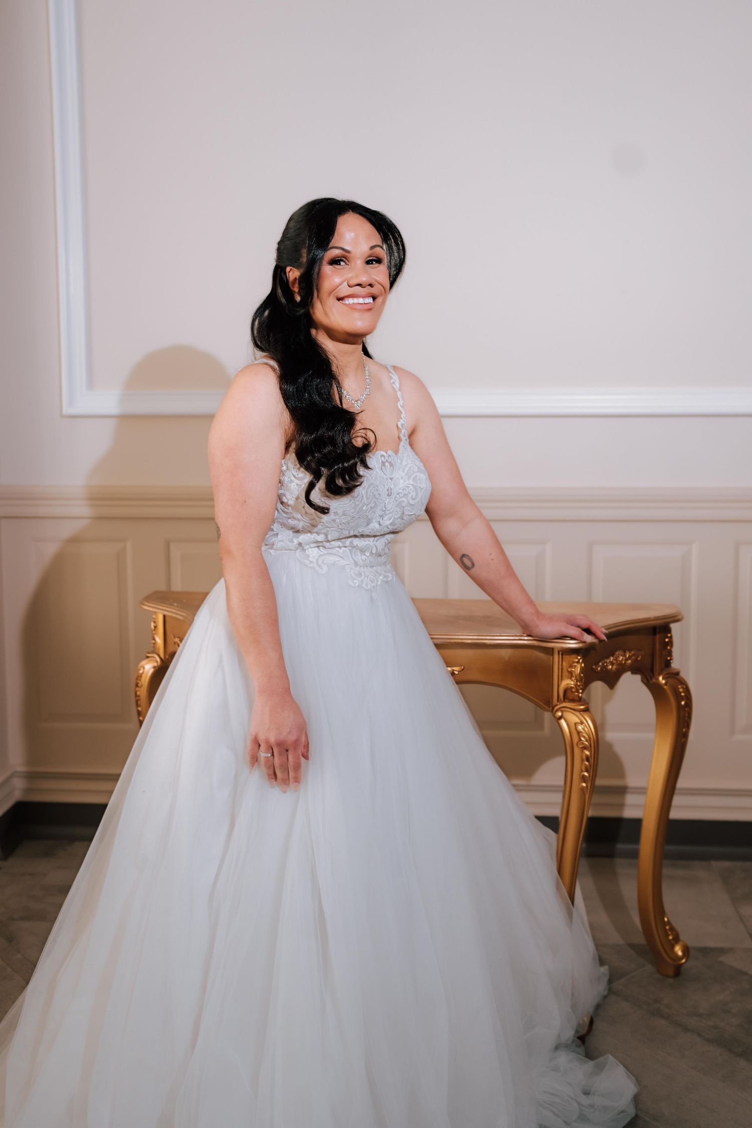 Beautiful bride in a white lace wedding dress smiling beside a gold antique table indoors.