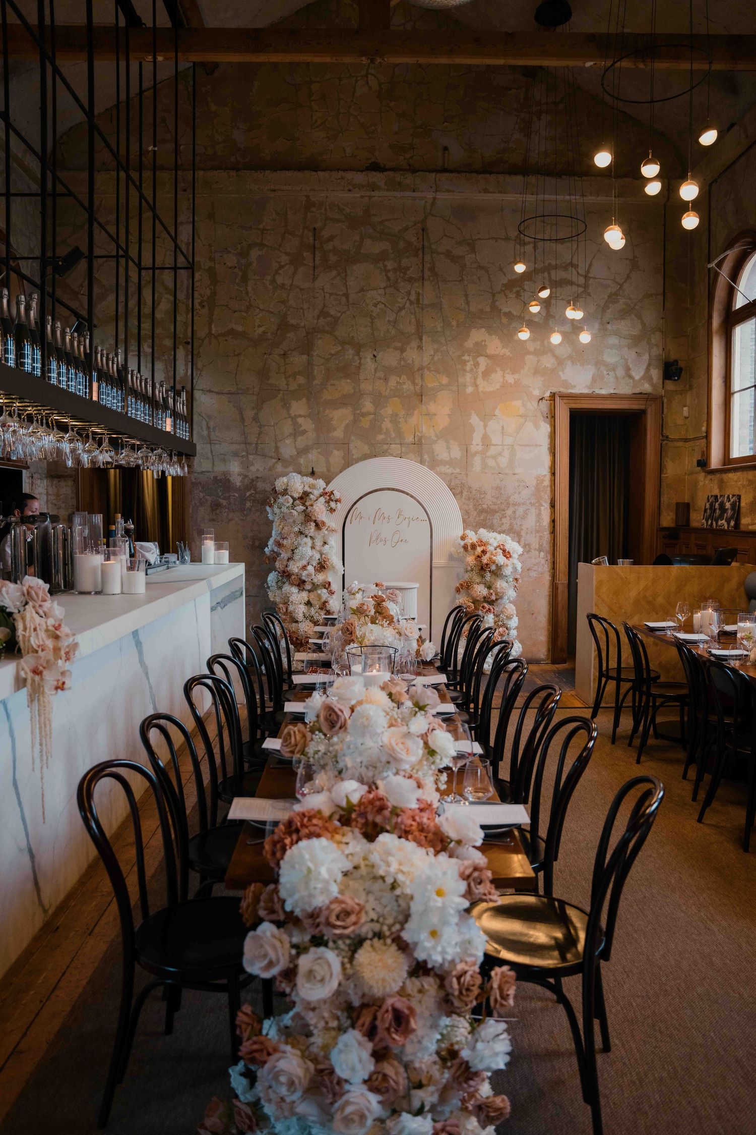 Elegant wedding reception table with lush blush floral runner, black bentwood chairs, and warm ambient lighting in stone venue.