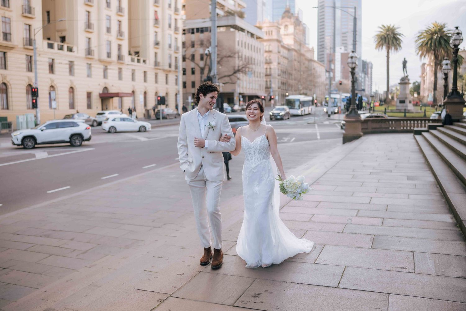 Smiling bride and groom walk together on city sidewalk with urban buildings in background.