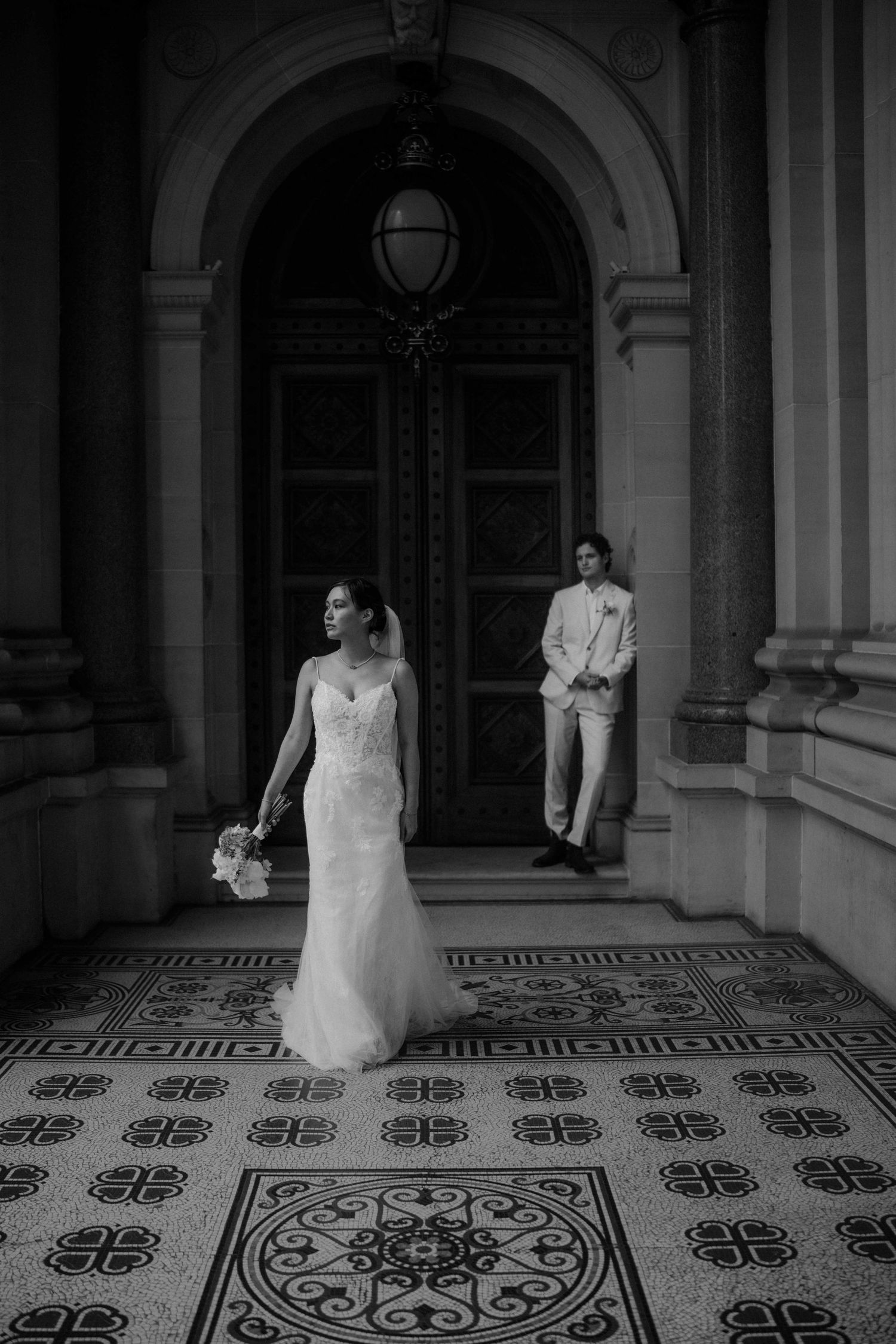 Black and white photo of elegant bride in lace gown and groom in beige suit at ornate doorway.