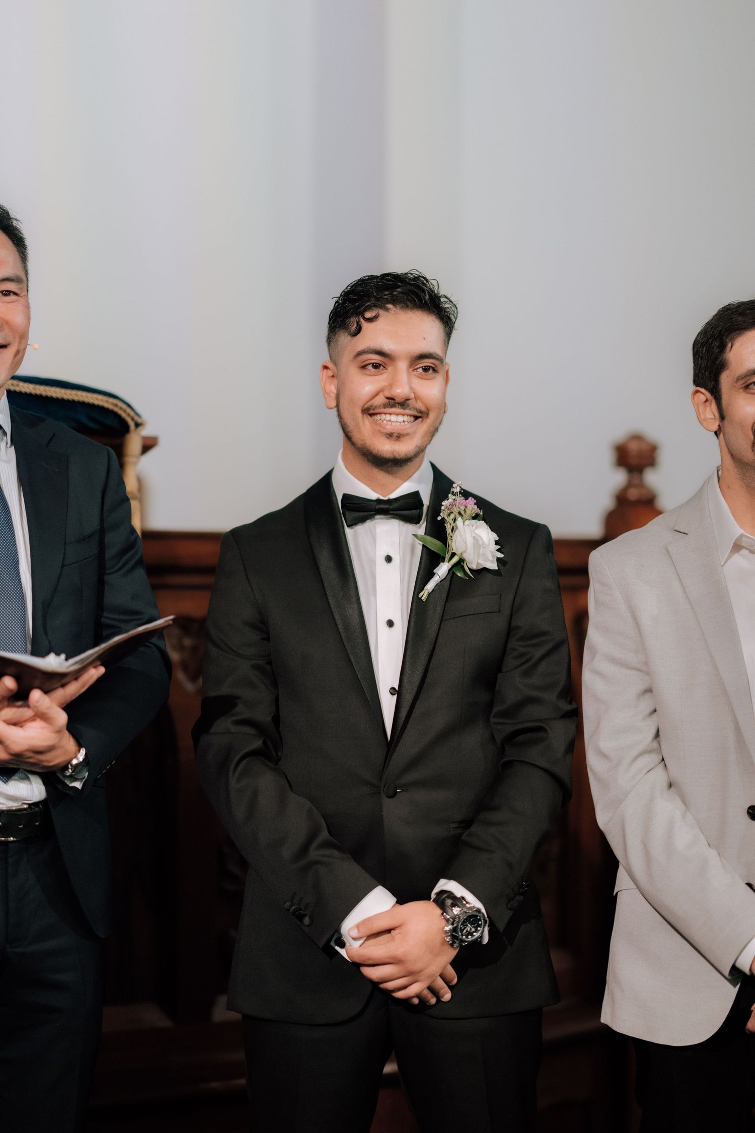 Groom in black tuxedo with boutonniere smiling at wedding ceremony altar.