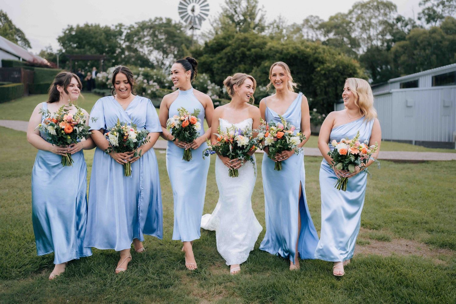 Bridal party of five bridesmaids in powder blue satin dresses walking with bride on green lawn.