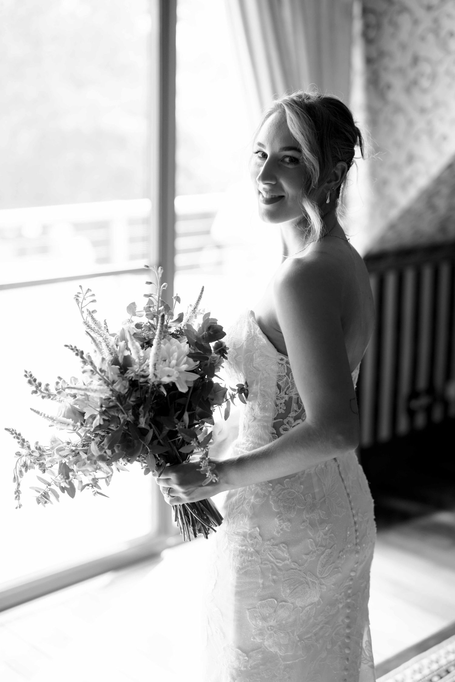 Black and white portrait of smiling bride holding floral bouquet by bright window.