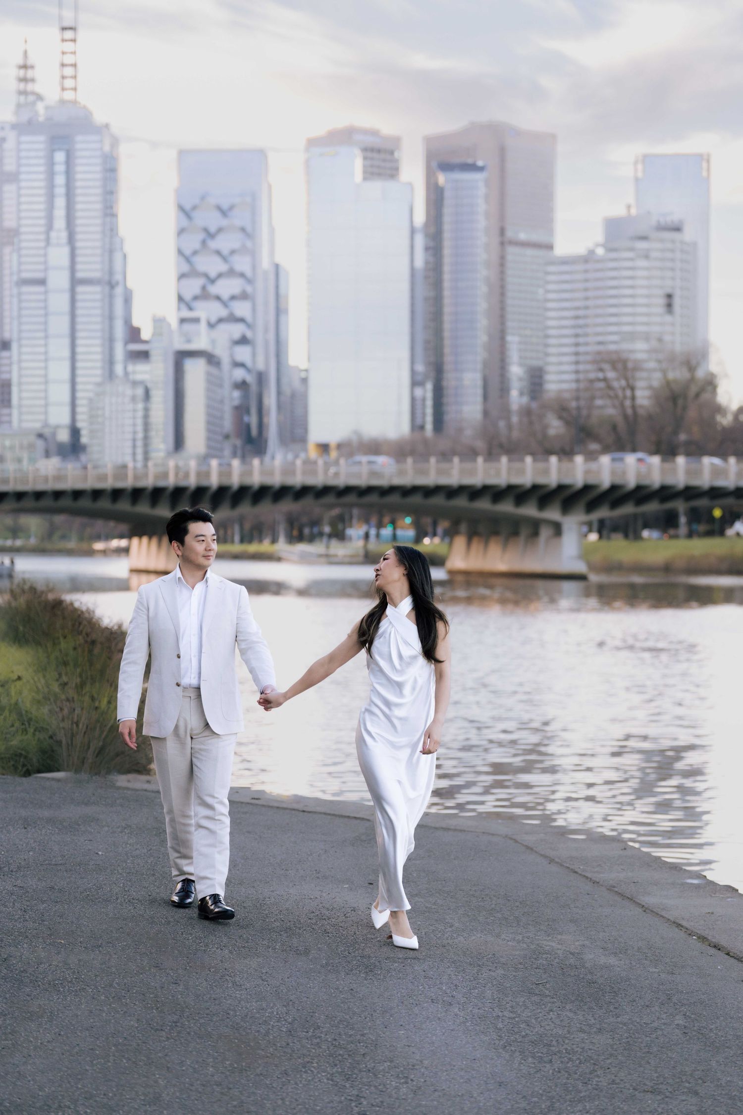 Couple in white outfits holding hands by a river with city skyline backdrop.