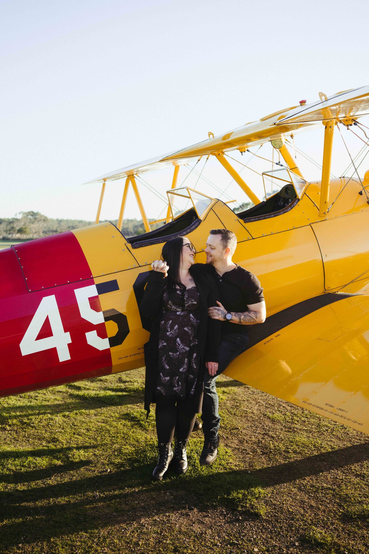 Couple embracing and smiling beside a vintage yellow biplane numbered 45 on a sunny airfield.