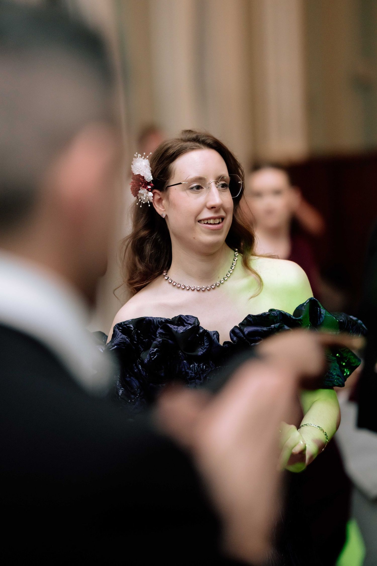 Woman in navy off-shoulder ruffled gown with floral hair accessory smiling at wedding reception.