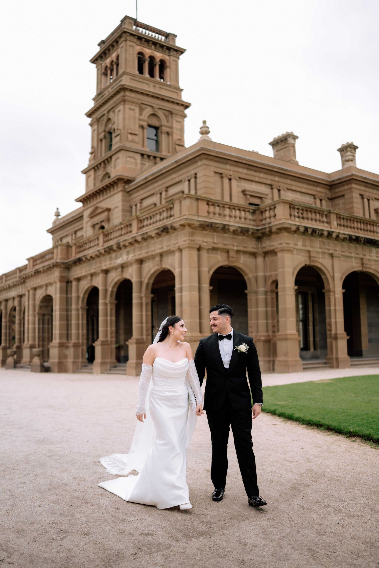 Bride and groom walk hand in hand before a grand historic sandstone building on their wedding day.