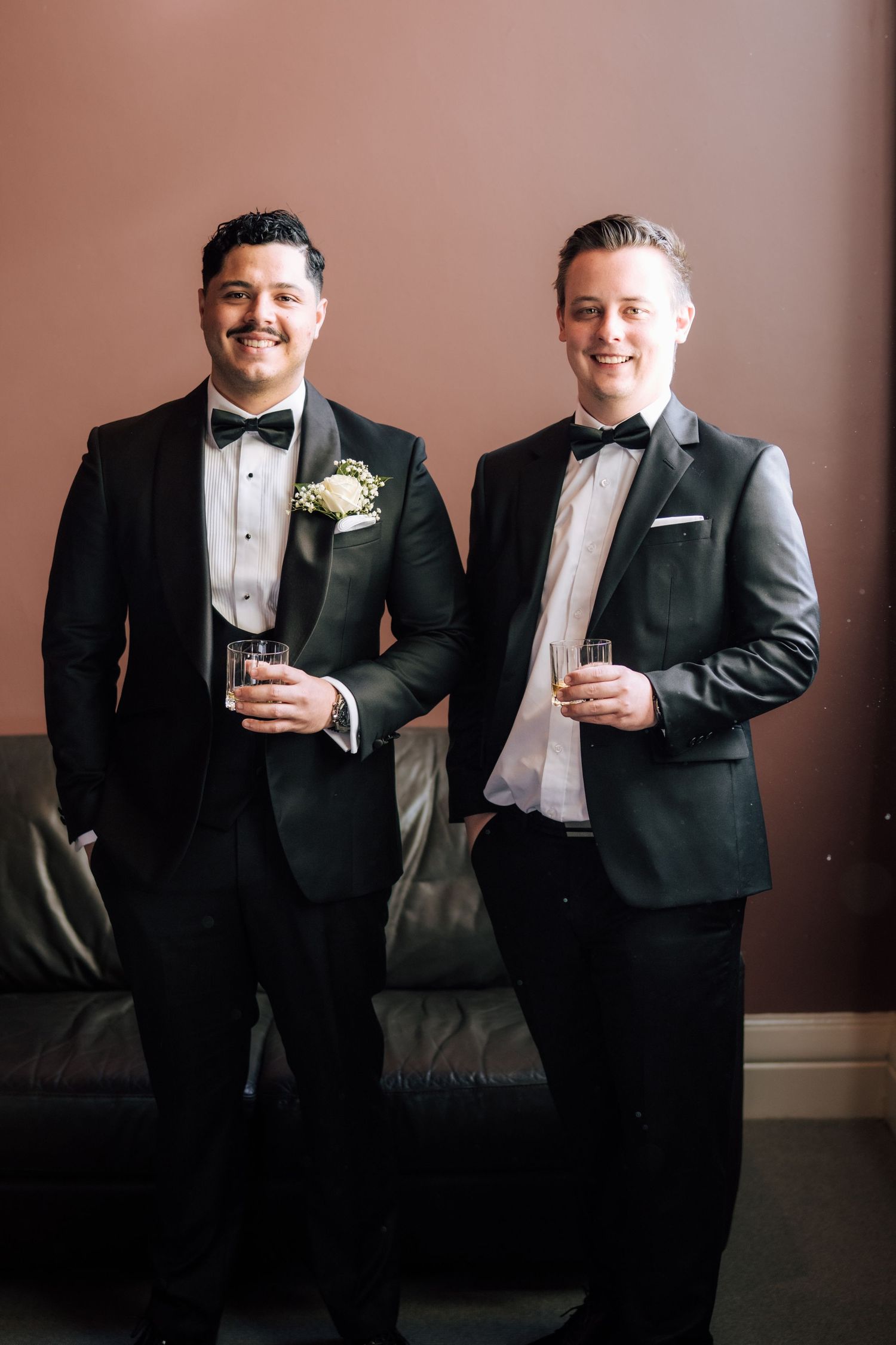Two grooms in black tuxedos with bow ties holding drinks and smiling on their wedding day.