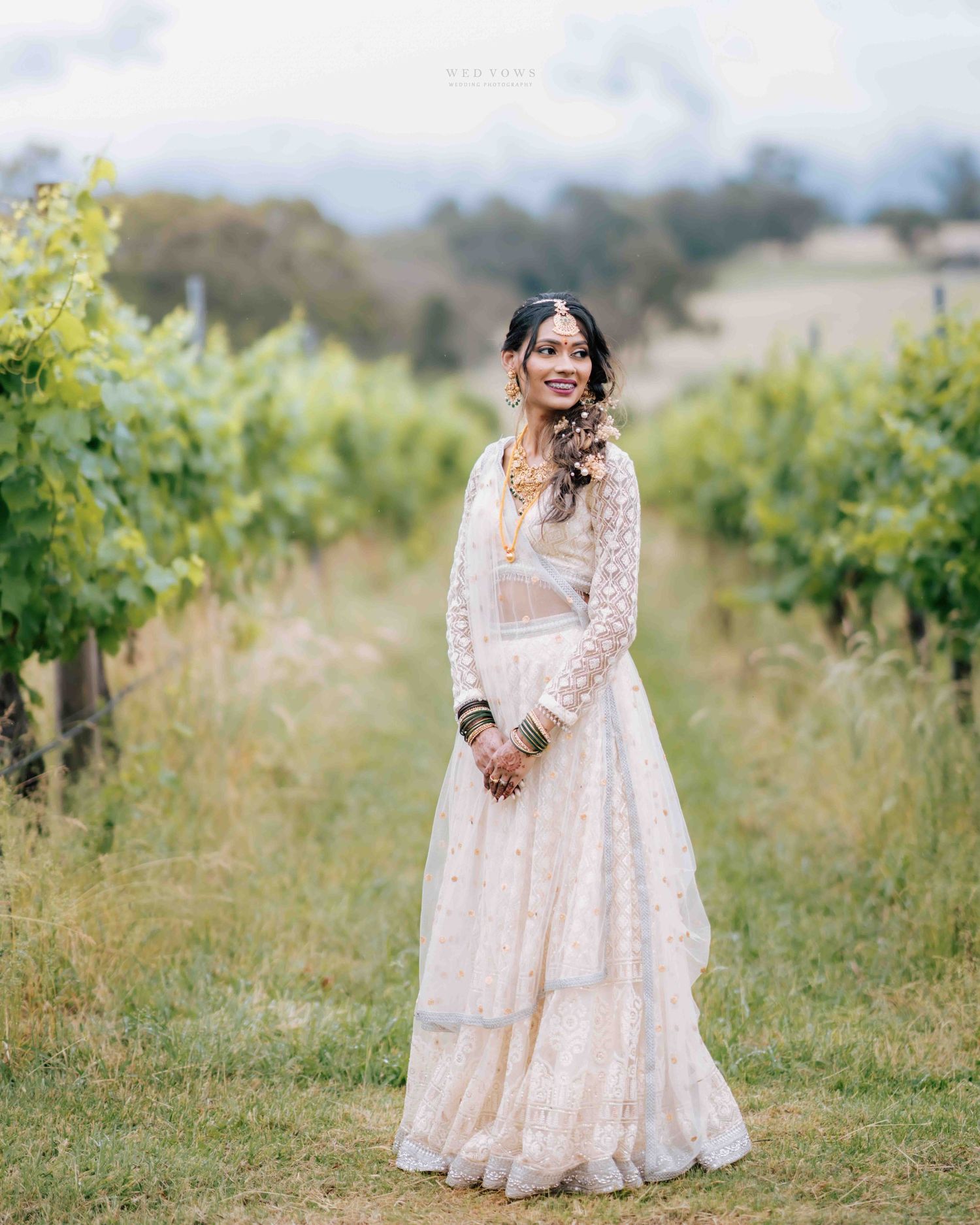 Indian bride in white embroidered lehenga smiling beautifully while standing between vineyard rows.