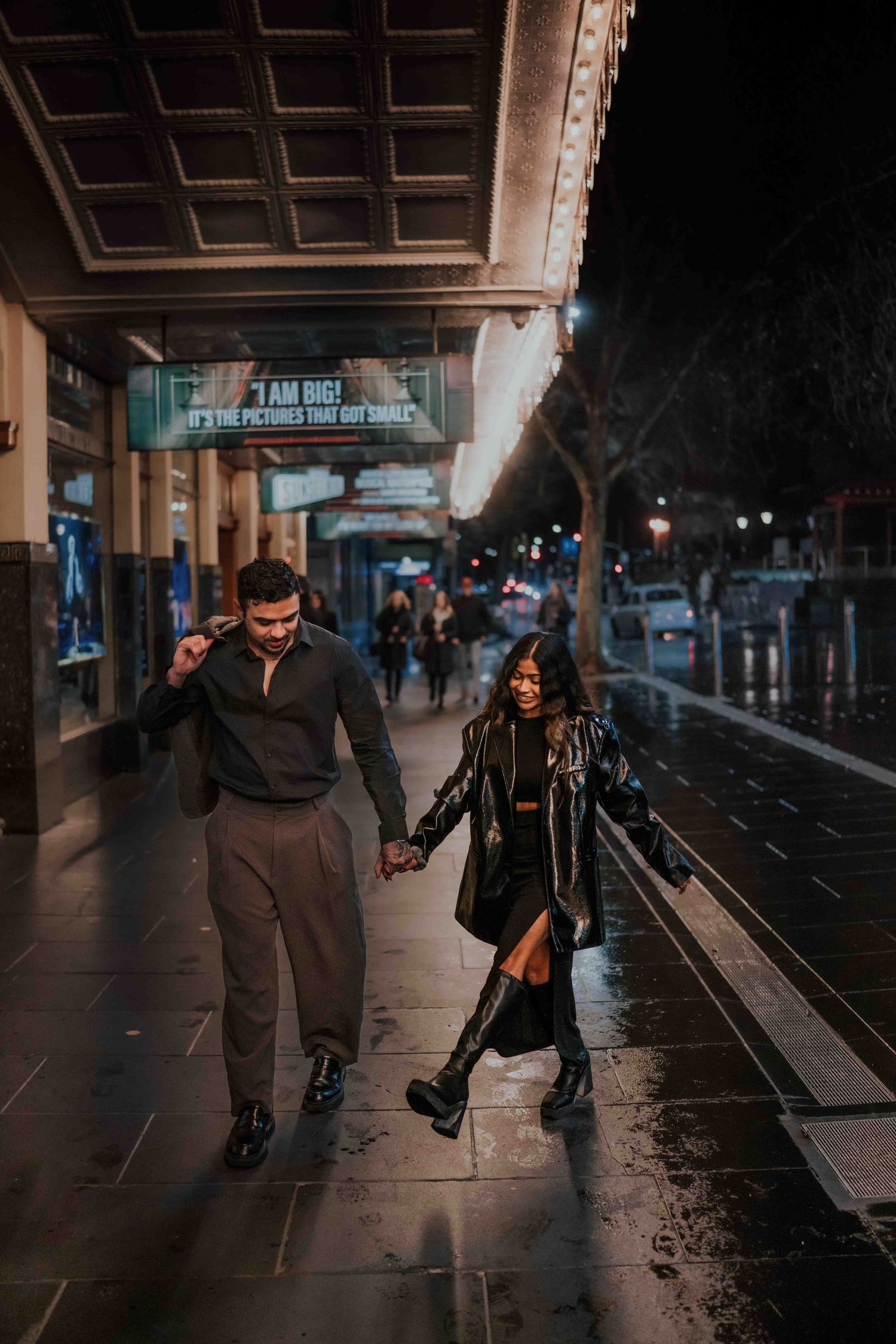 Couple strolling hand in hand on a wet sidewalk at night near a glowing theater entrance.