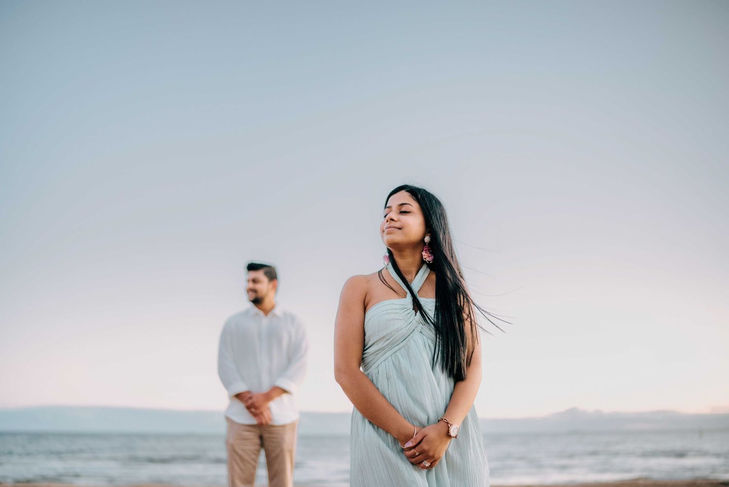 Woman in light blue halter dress standing on beach with man in white shirt blurred in background.