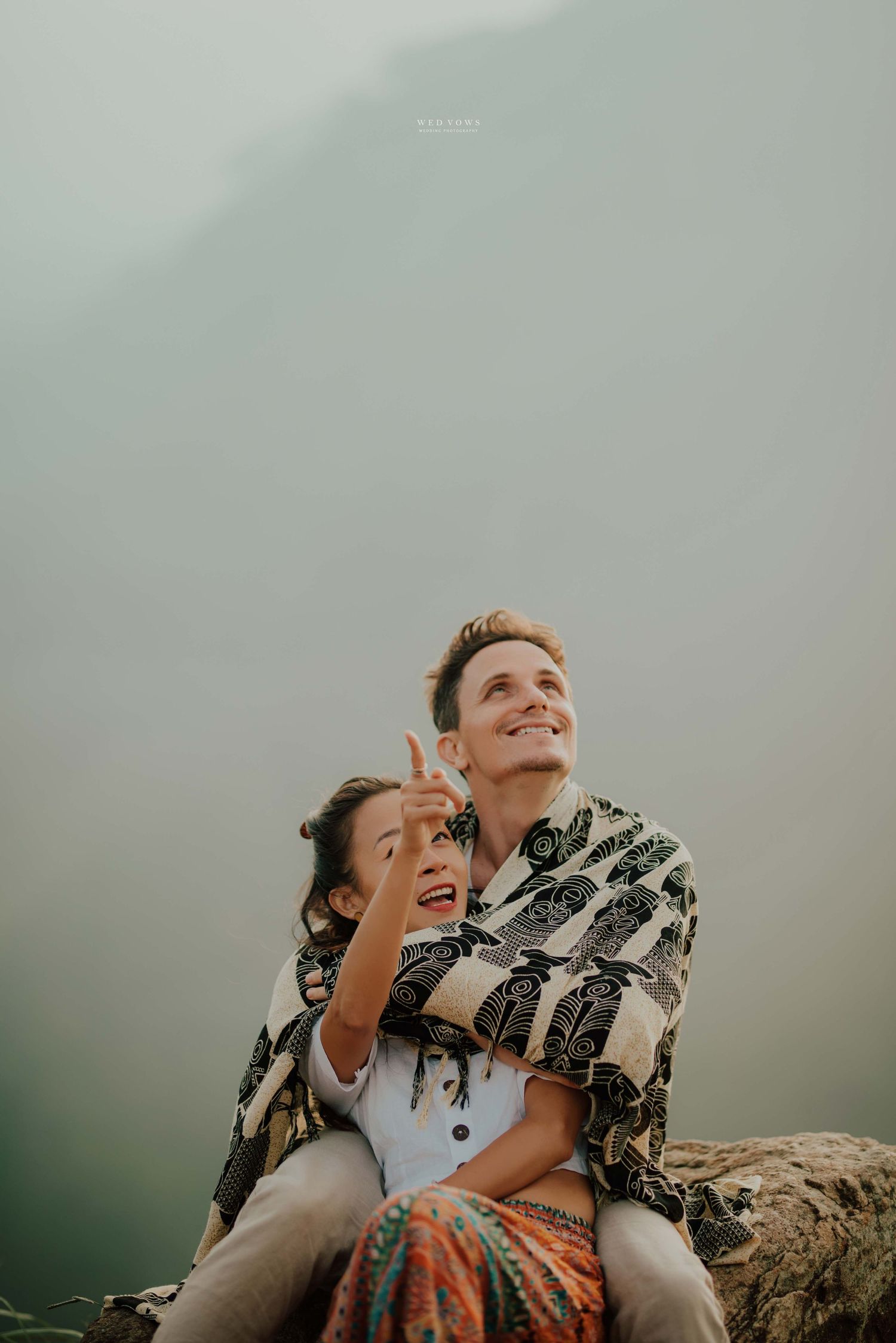 Smiling couple sitting on rocks pointing upward against misty mountain backdrop.