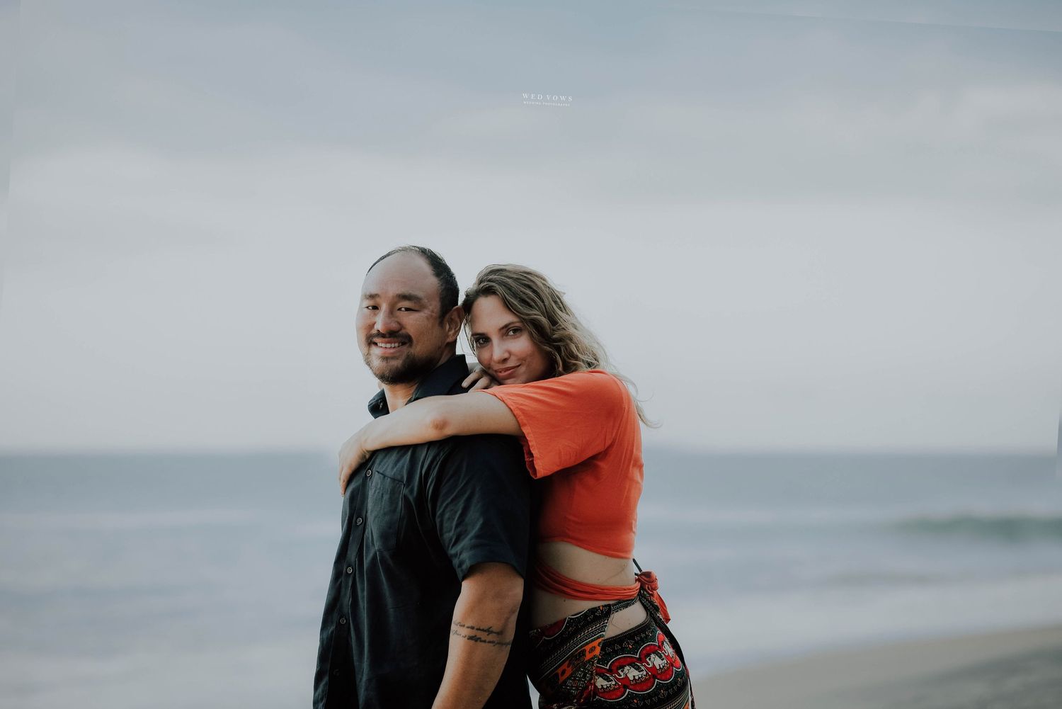 Couple embracing on a beach, woman hugging man from behind, both smiling with ocean in background.