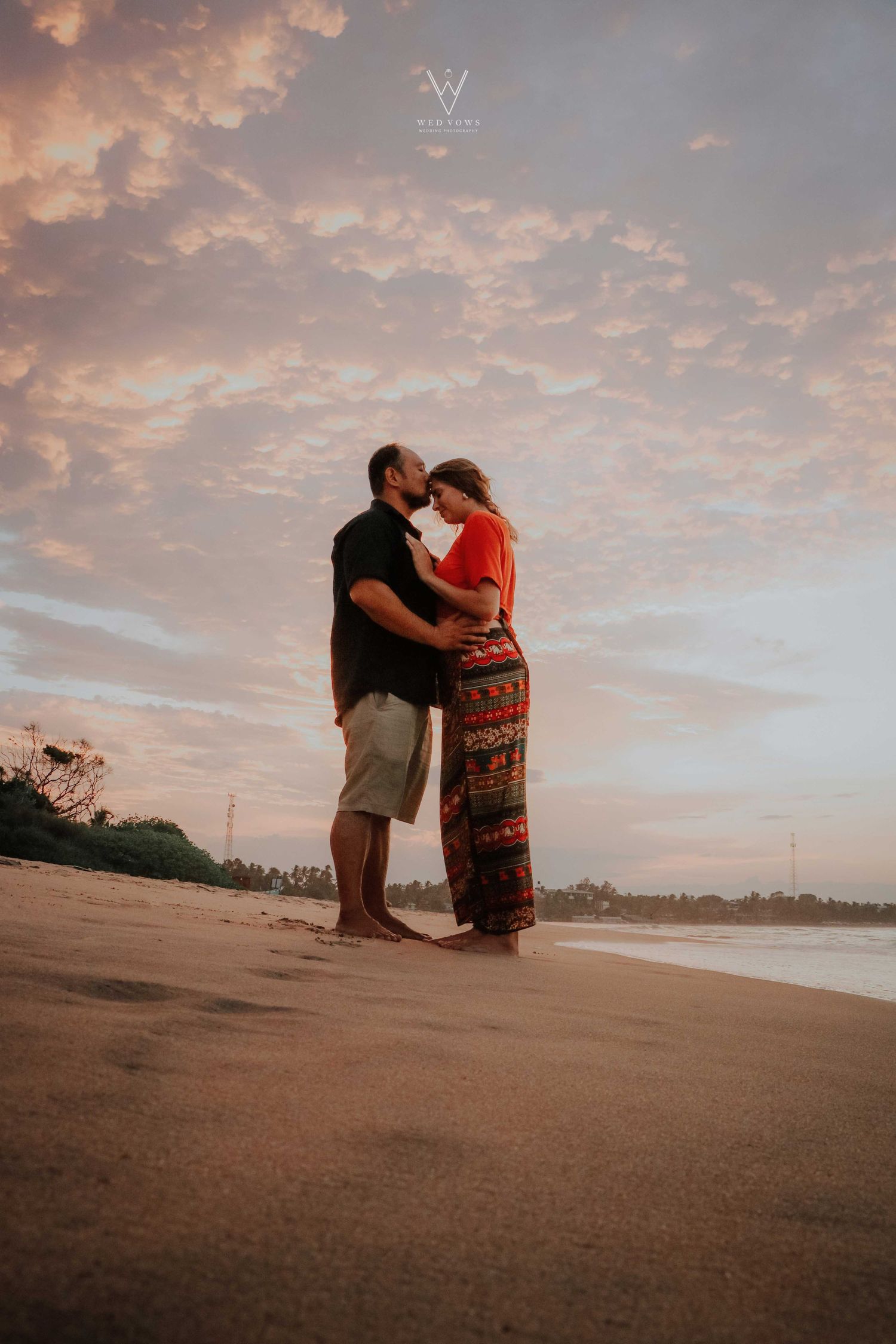 Couple sharing a romantic kiss on a tropical beach at sunset with dramatic pink cloudy sky.