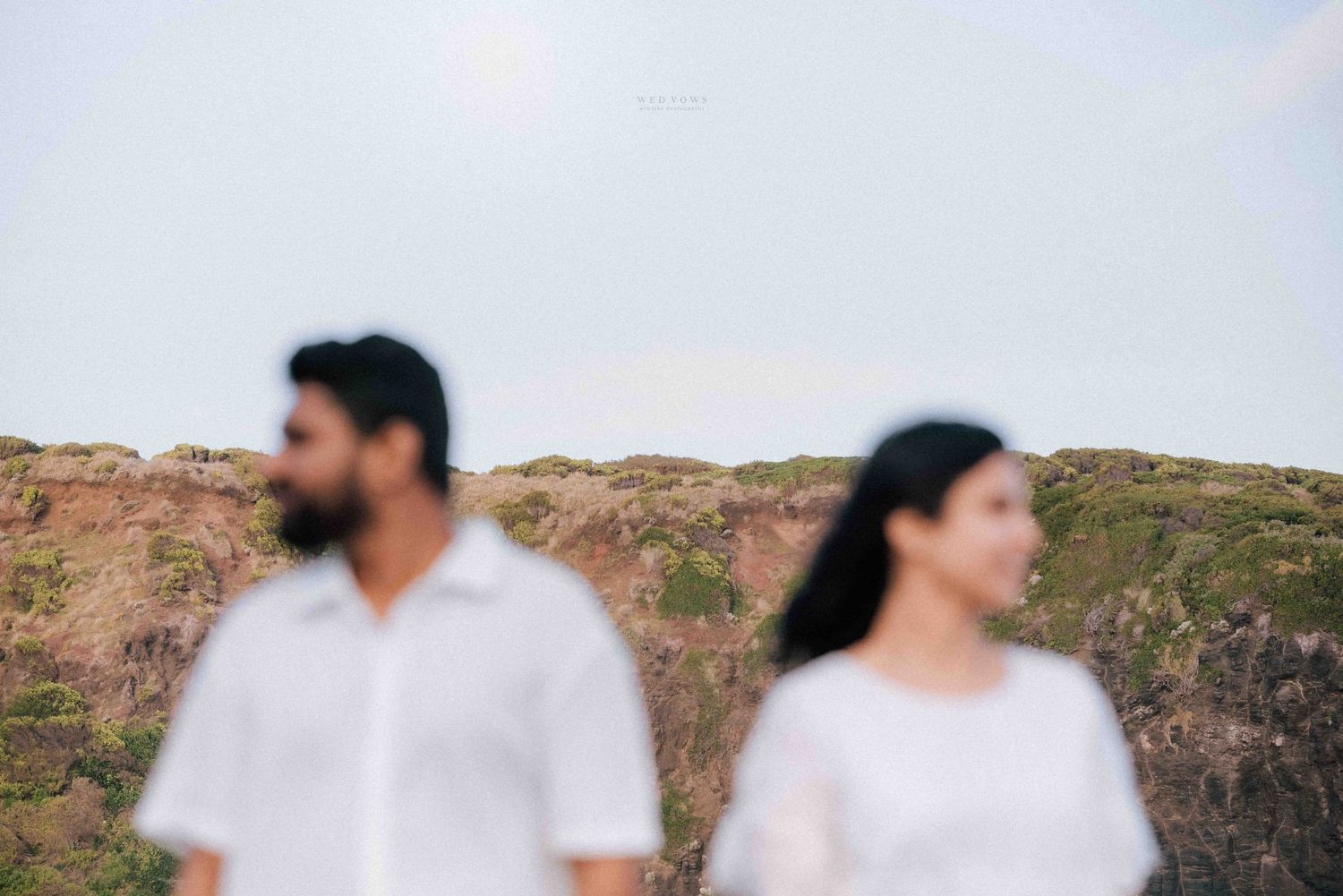 Couple in white outfits standing apart with dramatic rocky cliffs in the background.