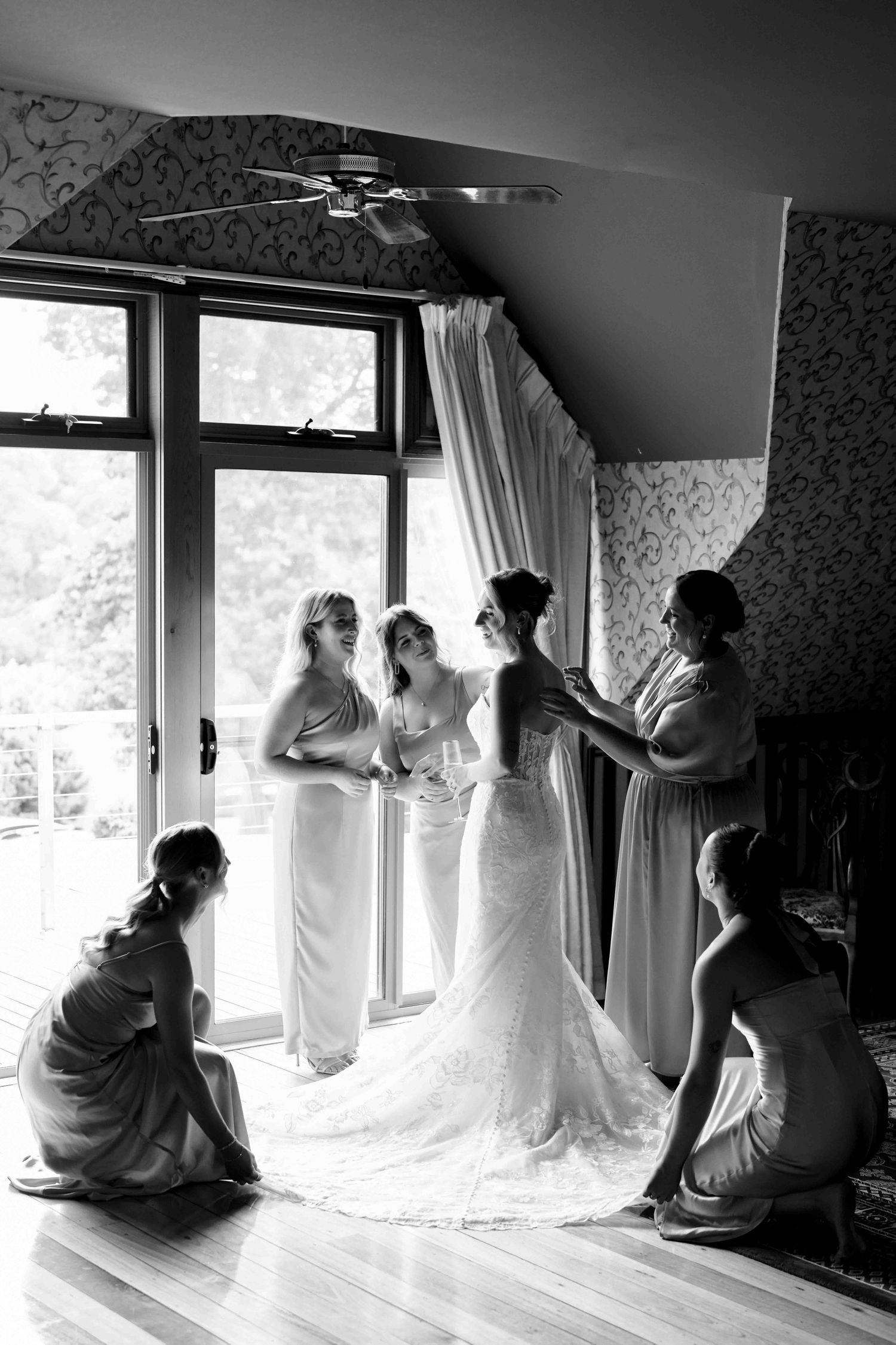 Black and white photo of bridesmaids helping bride into wedding dress near large windows with natural light.