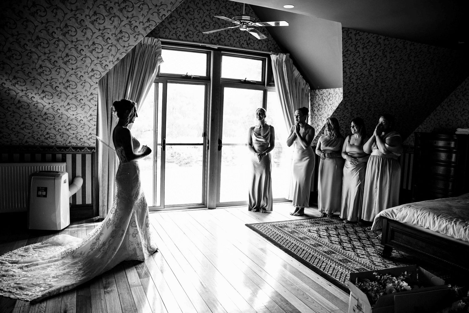 Black and white photo of bride in lace gown with bridesmaids cheering in elegant room.
