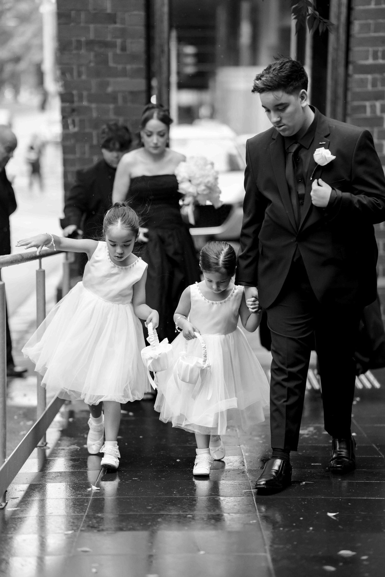 Young flower girls in white dresses walk with groomsman in black suit at wedding ceremony.
